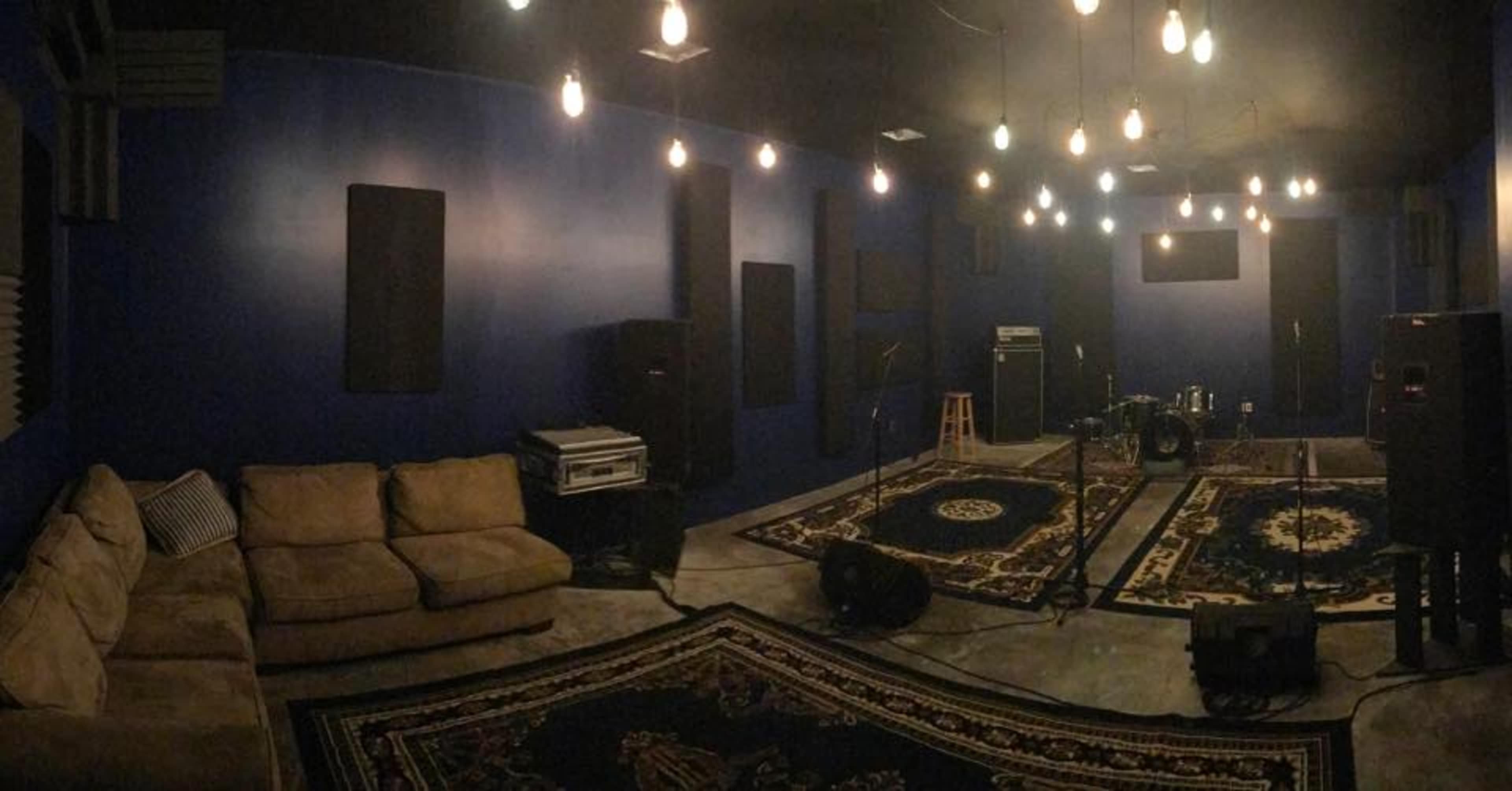 The image shows a dimly lit music rehearsal space with blue walls, several sound equipment setups, and a couch arranged on a patterned rug.