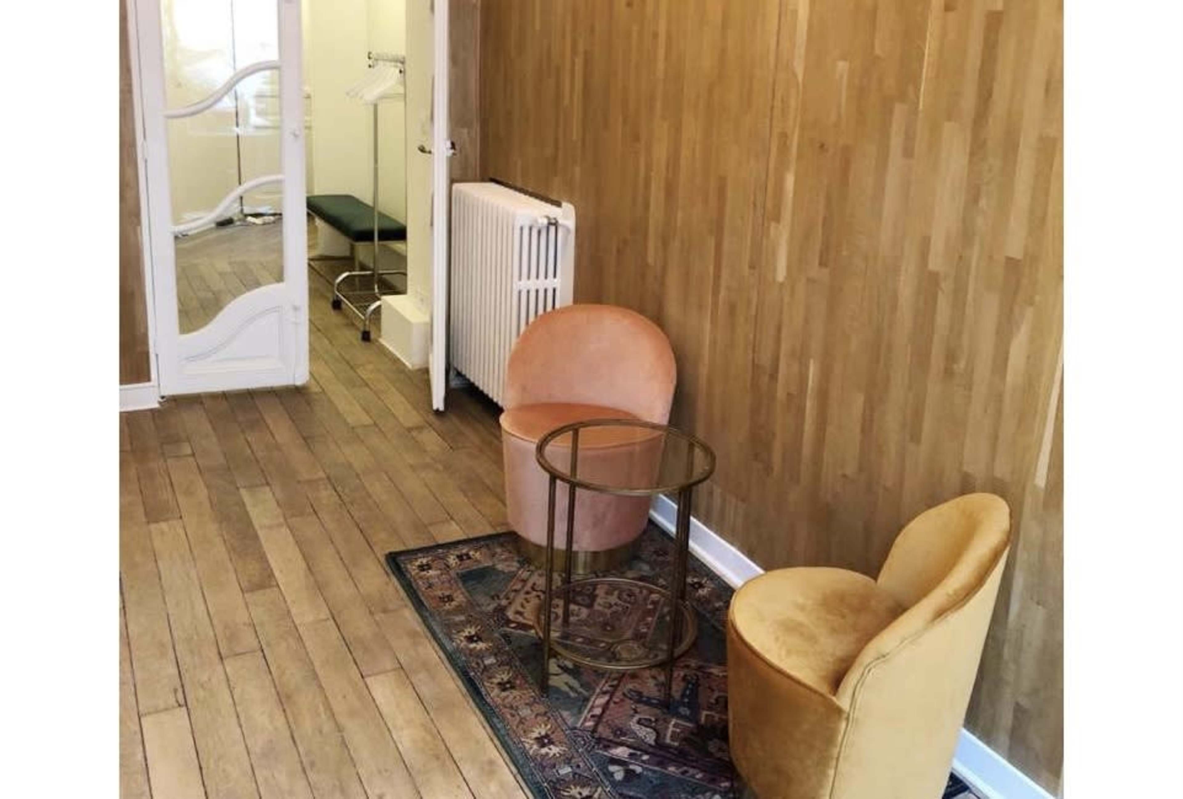 A small room with two upholstered chairs, a round glass table, and a mirror reflecting a massage bed and a radiator.