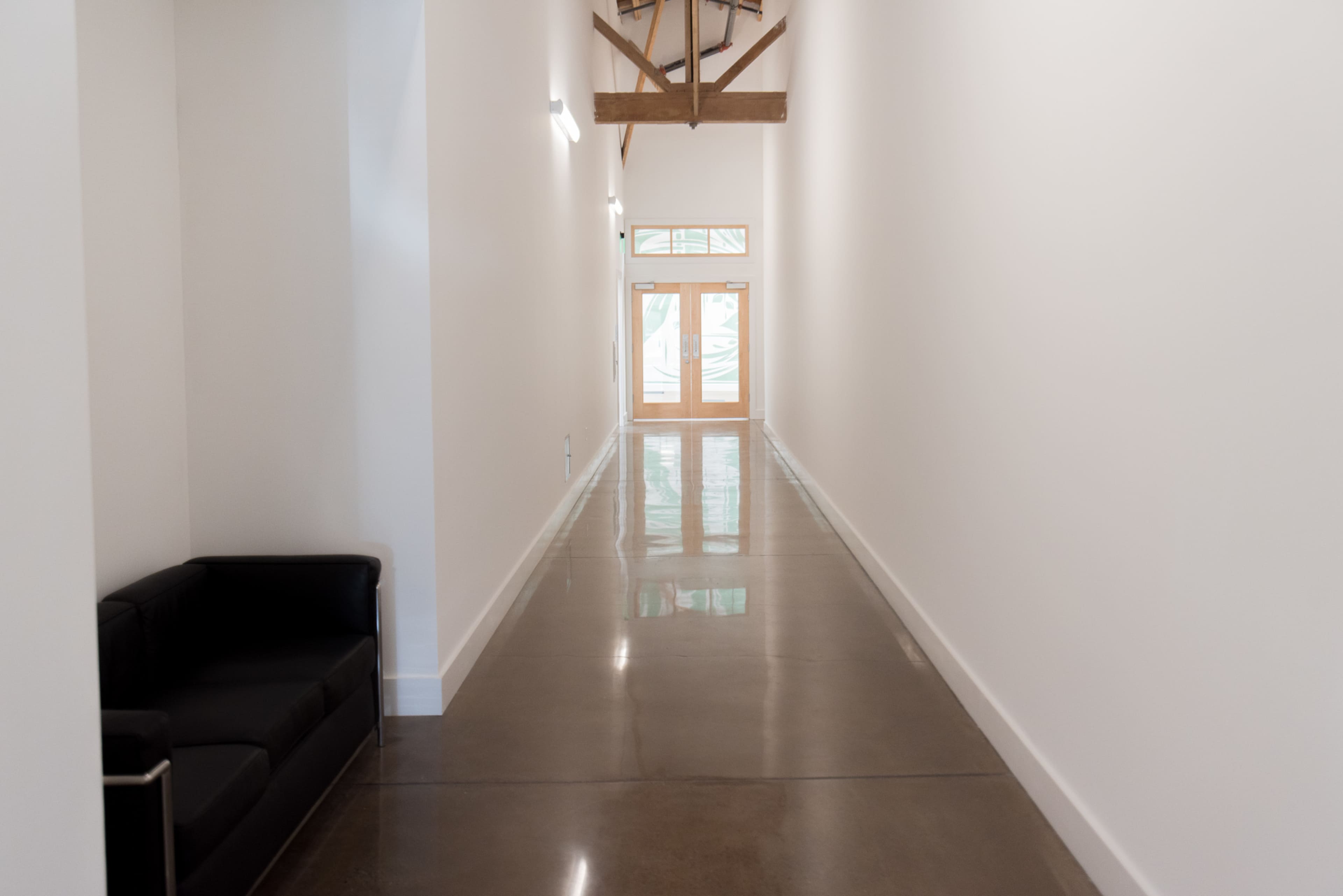 A long, narrow hallway features a polished concrete floor, white walls, and a black couch against one side, leading to a set of glass doors at the end.