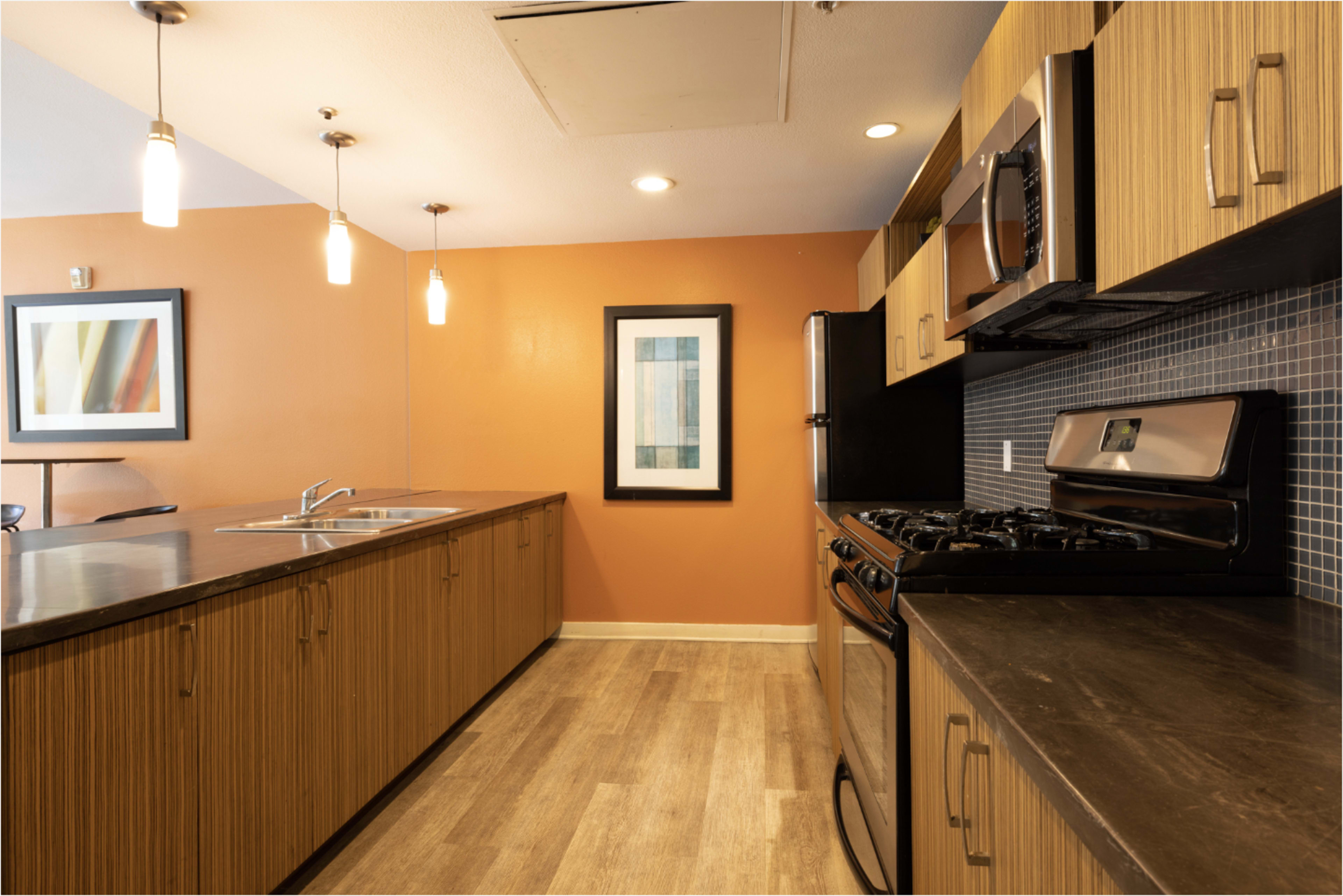 The image shows a modern kitchen with wooden cabinetry, stainless steel appliances, and an orange accent wall.