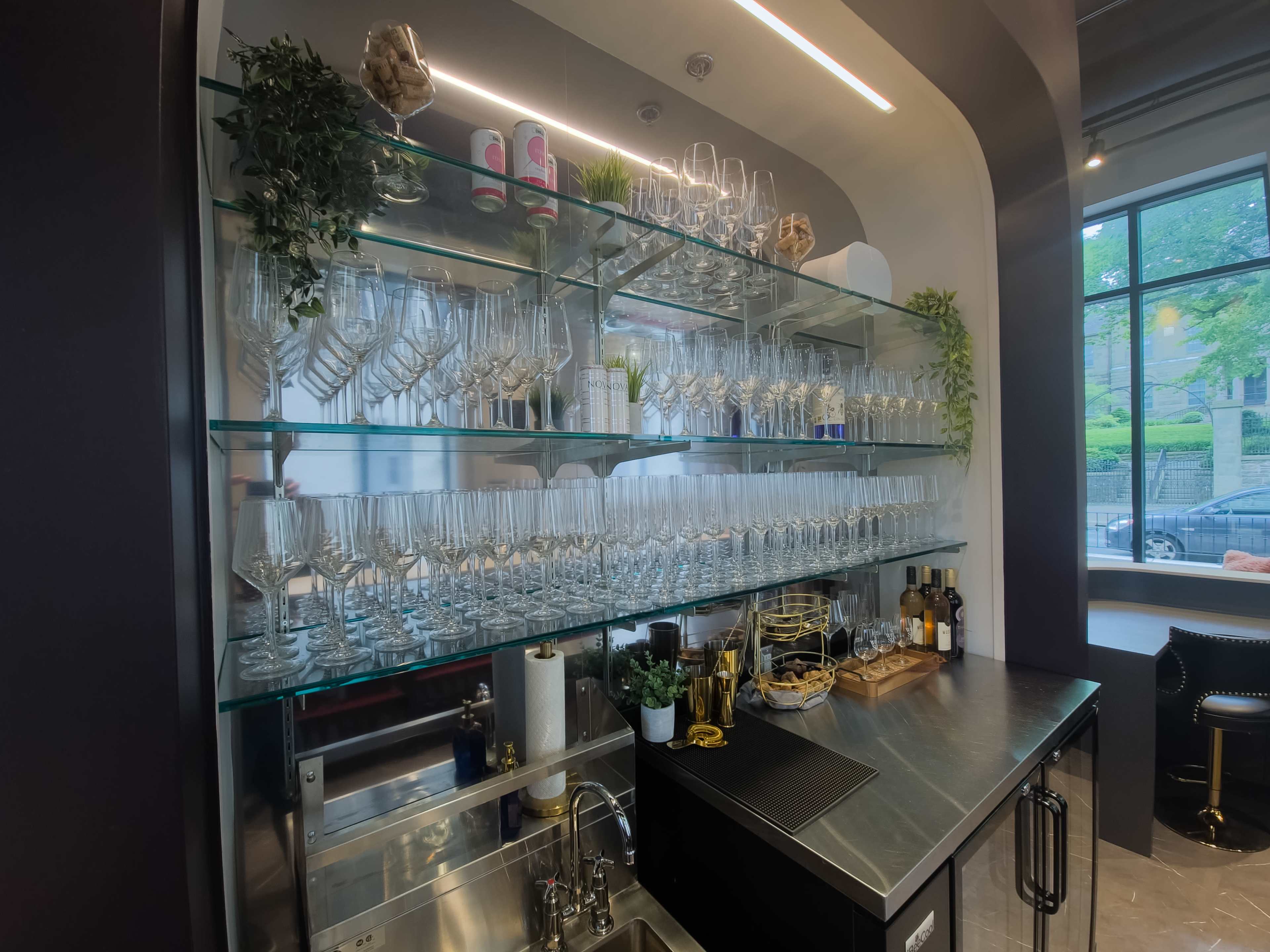 A modern bar area features a glass shelf filled with neatly arranged wine glasses and various bottles on a counter below.