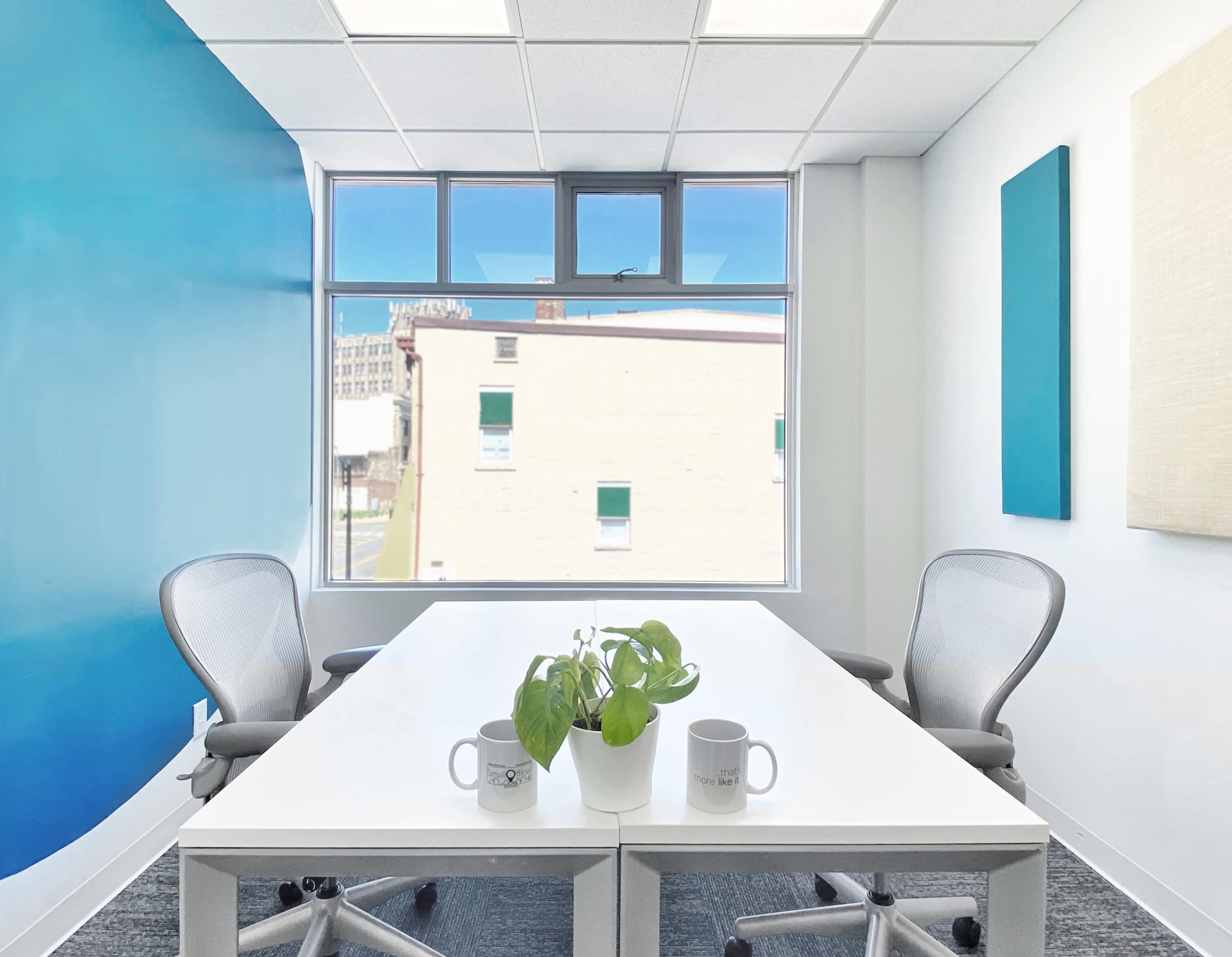 A bright office meeting room features a white table with two chairs, a small plant, and a large window overlooking a city scene.