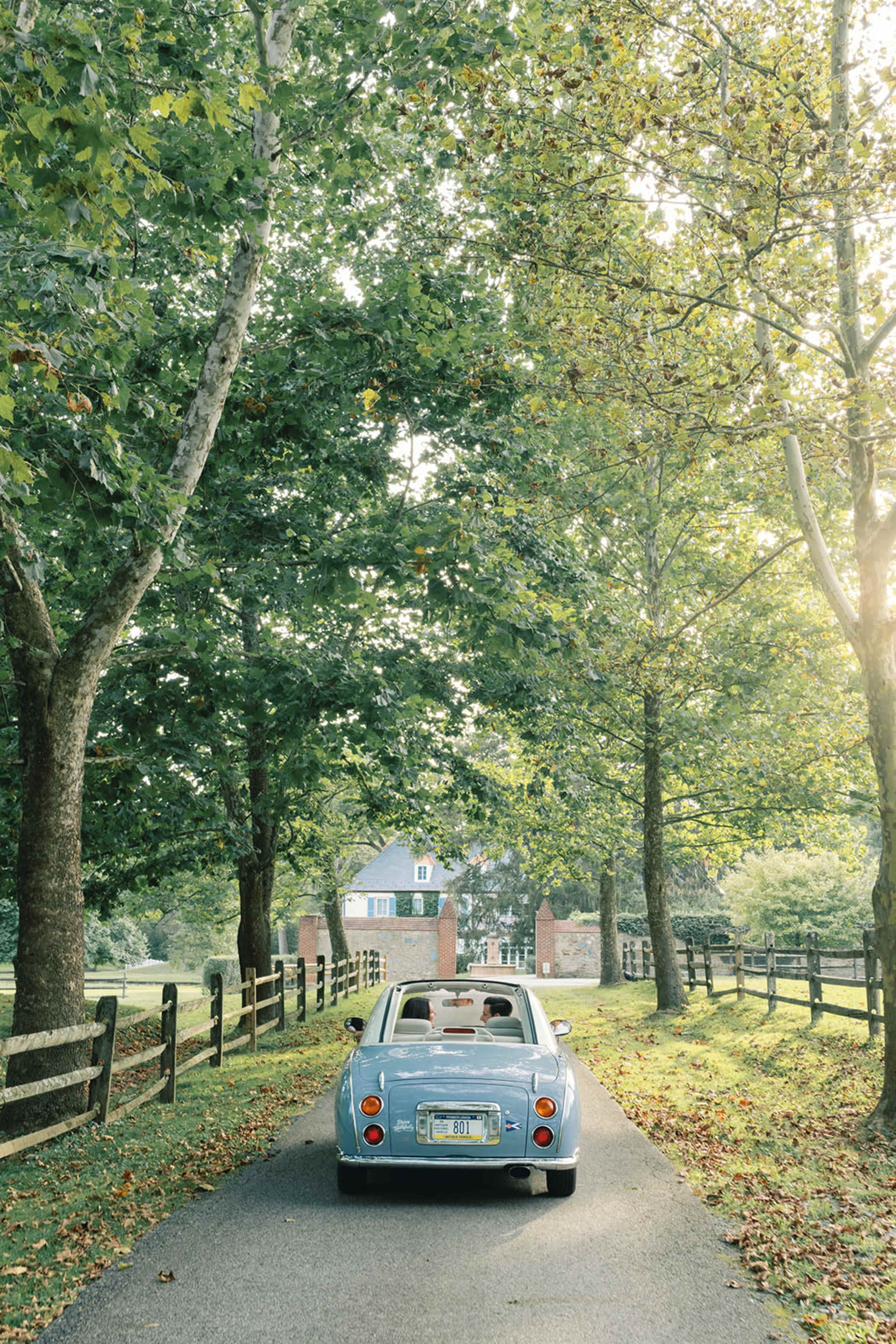 A vintage blue car is parked on a tree-lined road leading to a house in the distance.