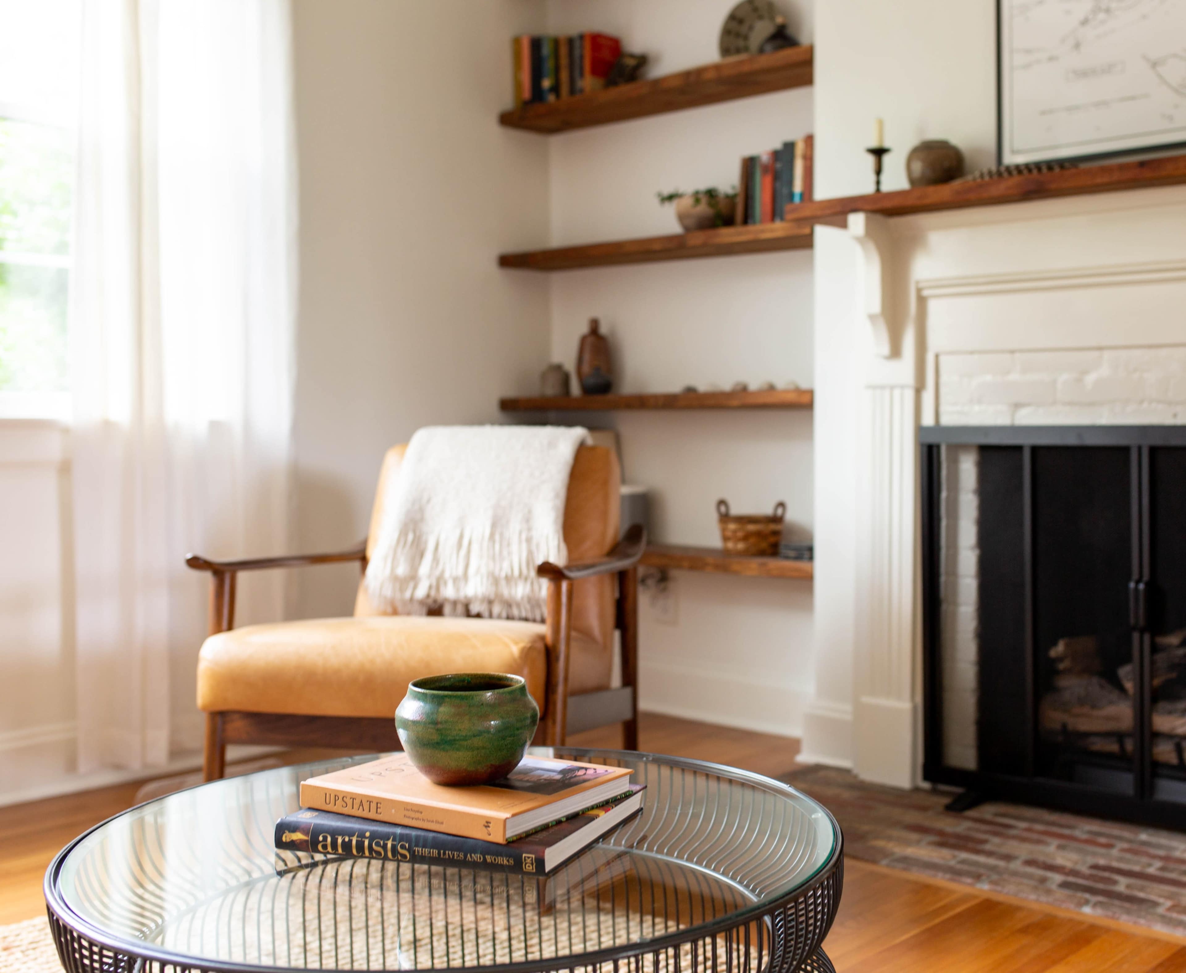 A living room features a glass coffee table with stacked books and a ceramic bowl, an armchair, and a shelf with decorative items.