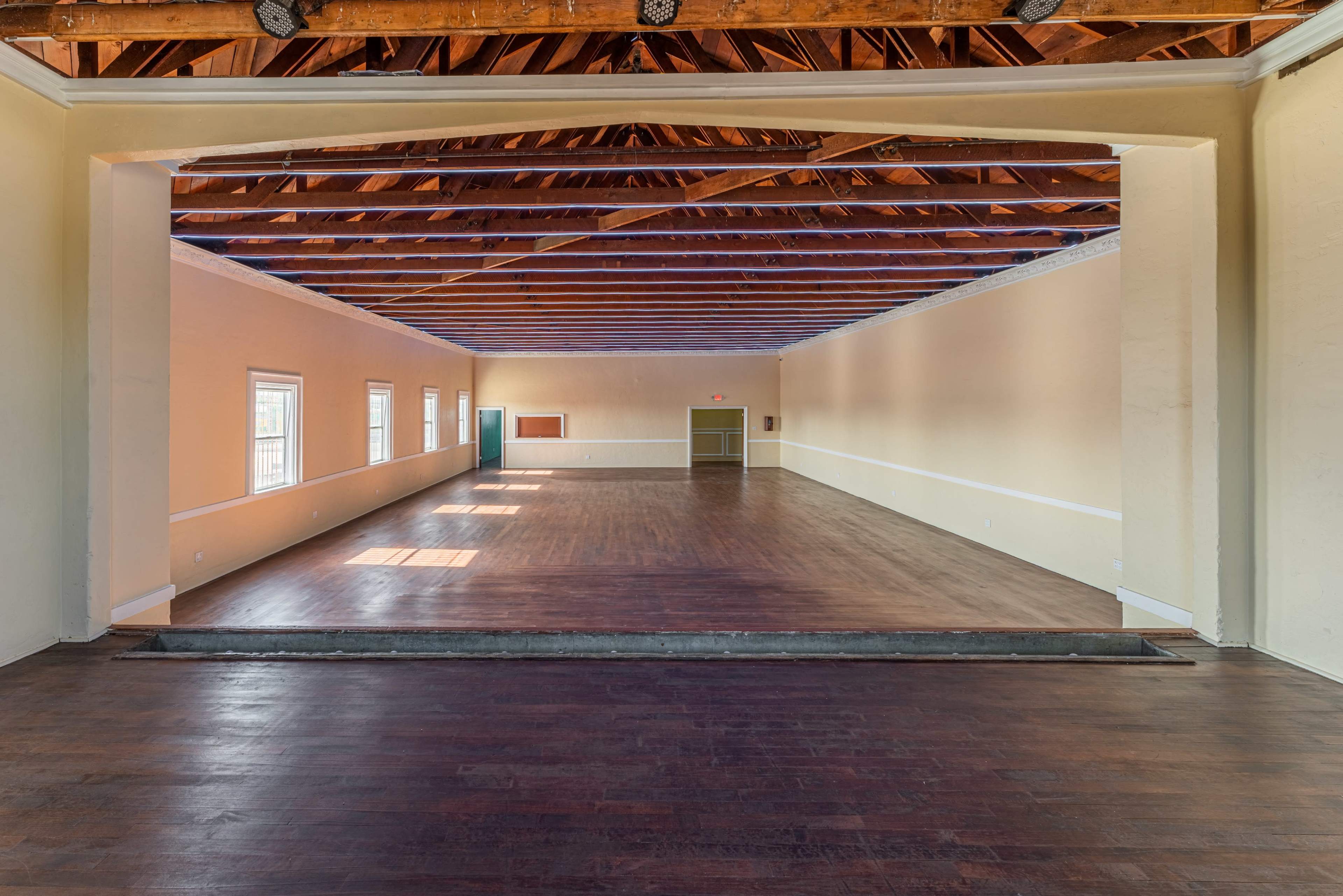 A spacious interior room with a wooden floor, high ceilings, and large windows on one side.