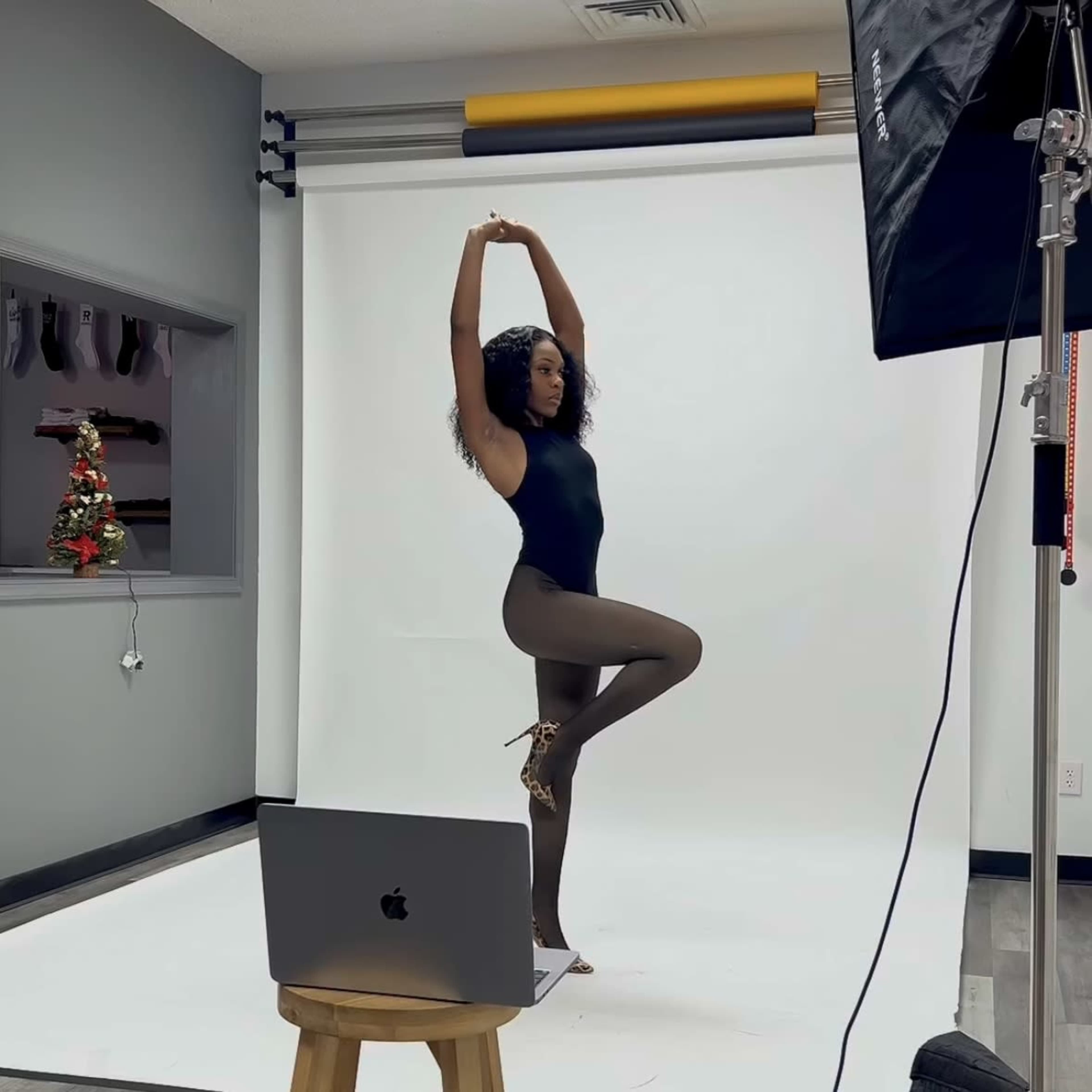 A dancer in a black leotard and sheer tights poses gracefully in a photography studio with a backdrop and a laptop on a stool.