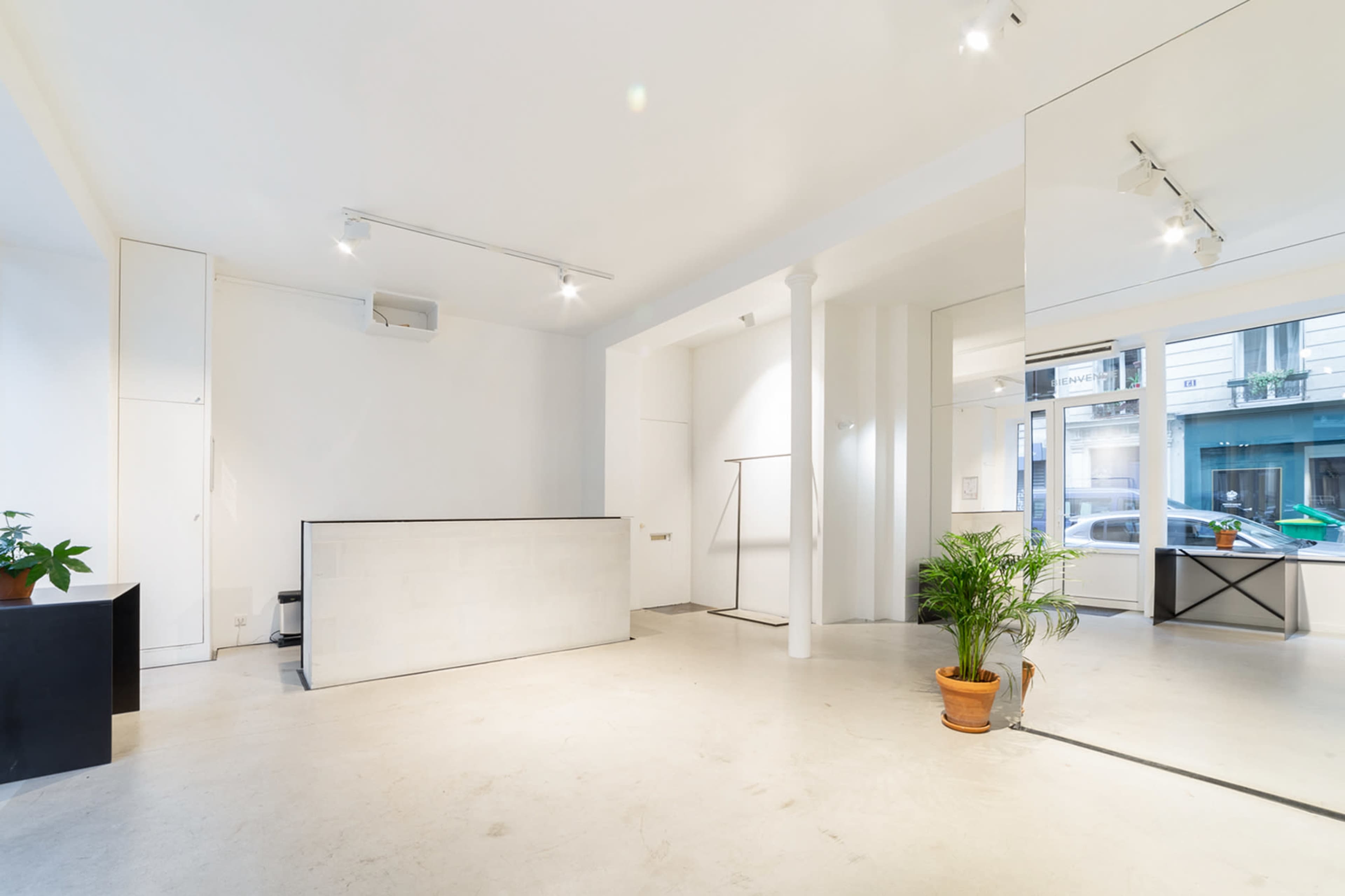 The image depicts a minimalist indoor space featuring a reception counter, a large window with a city view, and a potted plant.