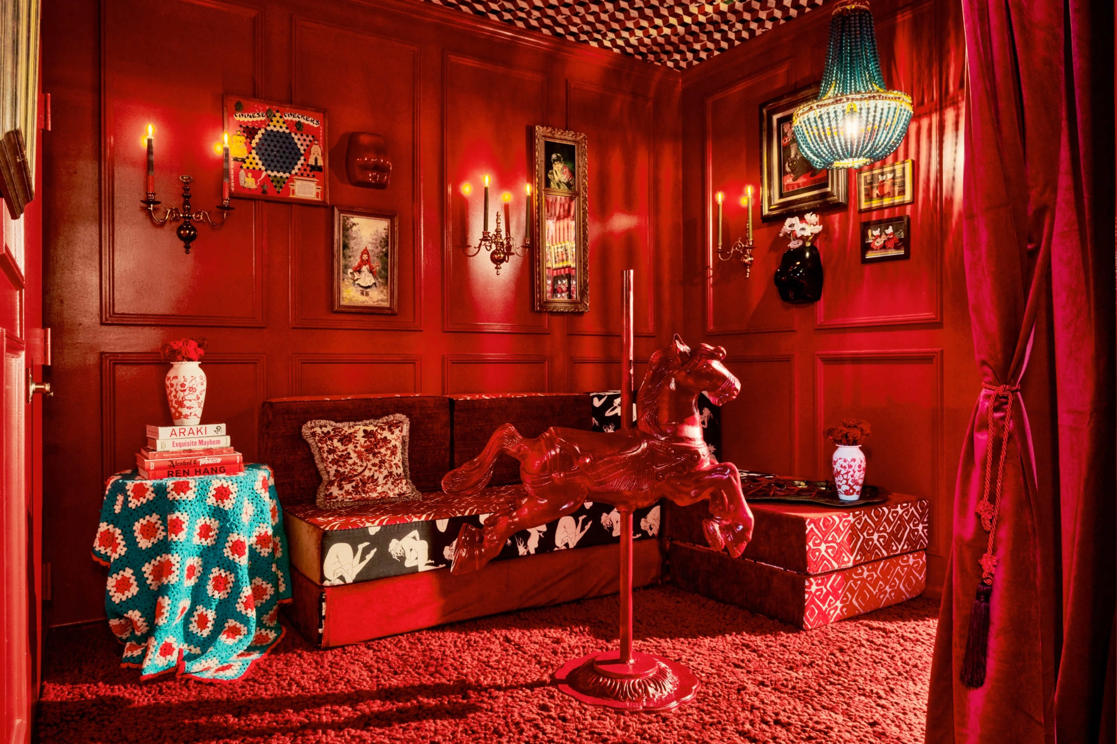 A small, dimly lit room with red walls, a patterned ceiling, a vintage rocking horse, and eclectic decor including a couch and various artwork.
