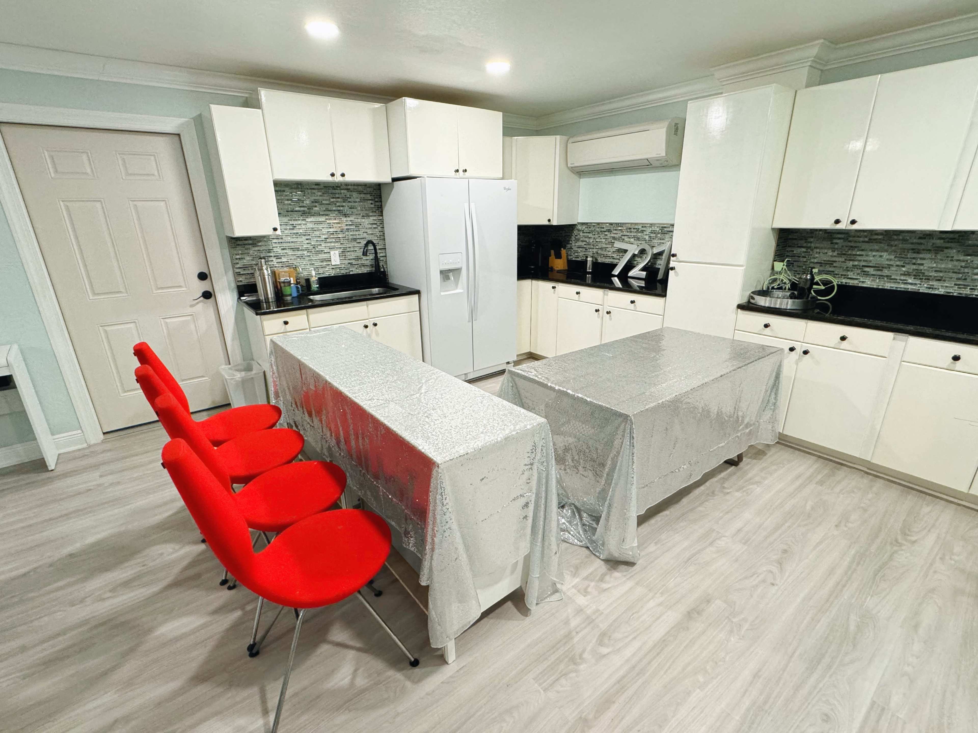 A modern kitchen features white cabinets, black countertops, and two silver-covered tables with red chairs.
