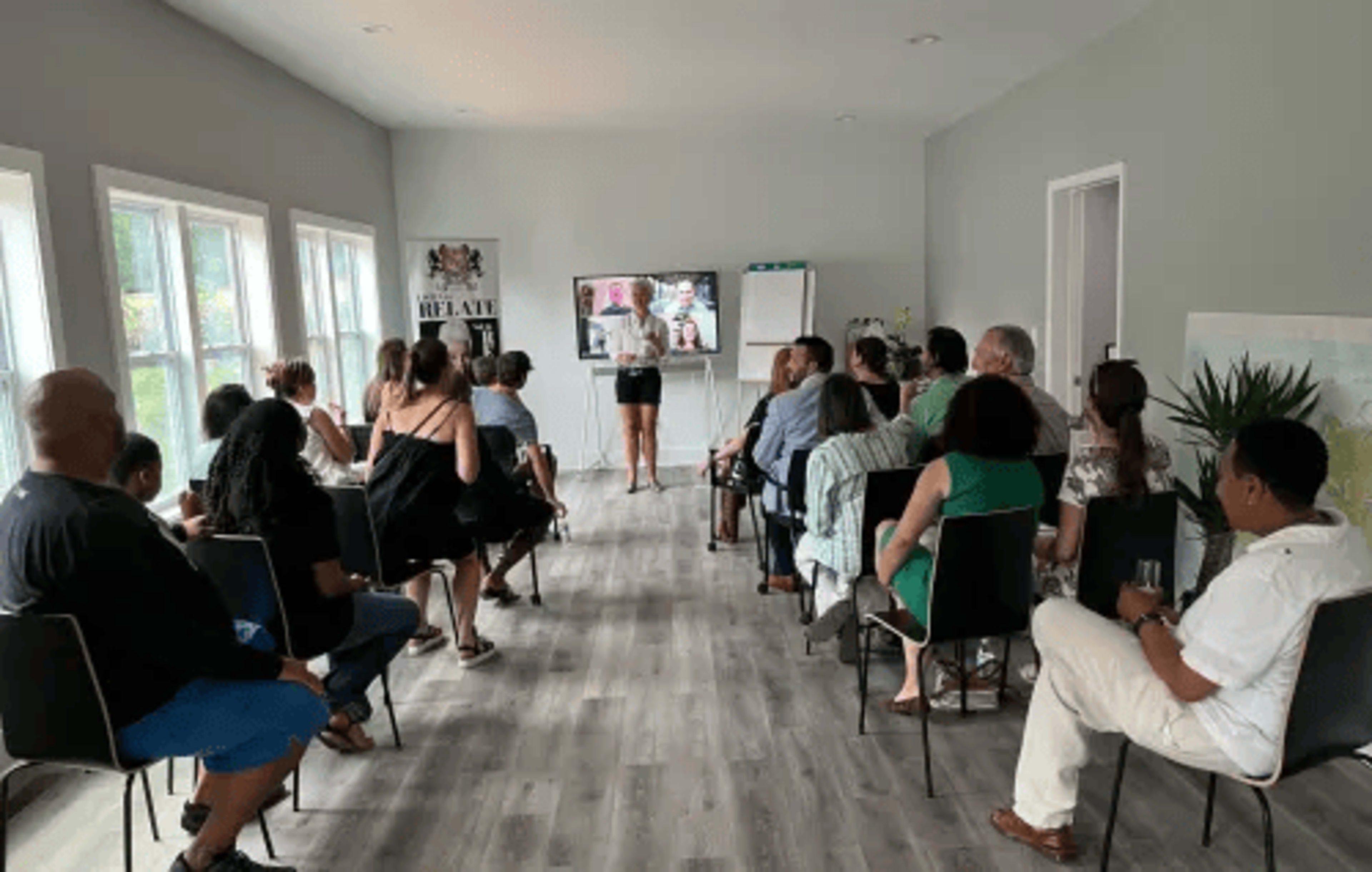 A group of people sits facing a woman presenting in front of a screen in a well-lit room.