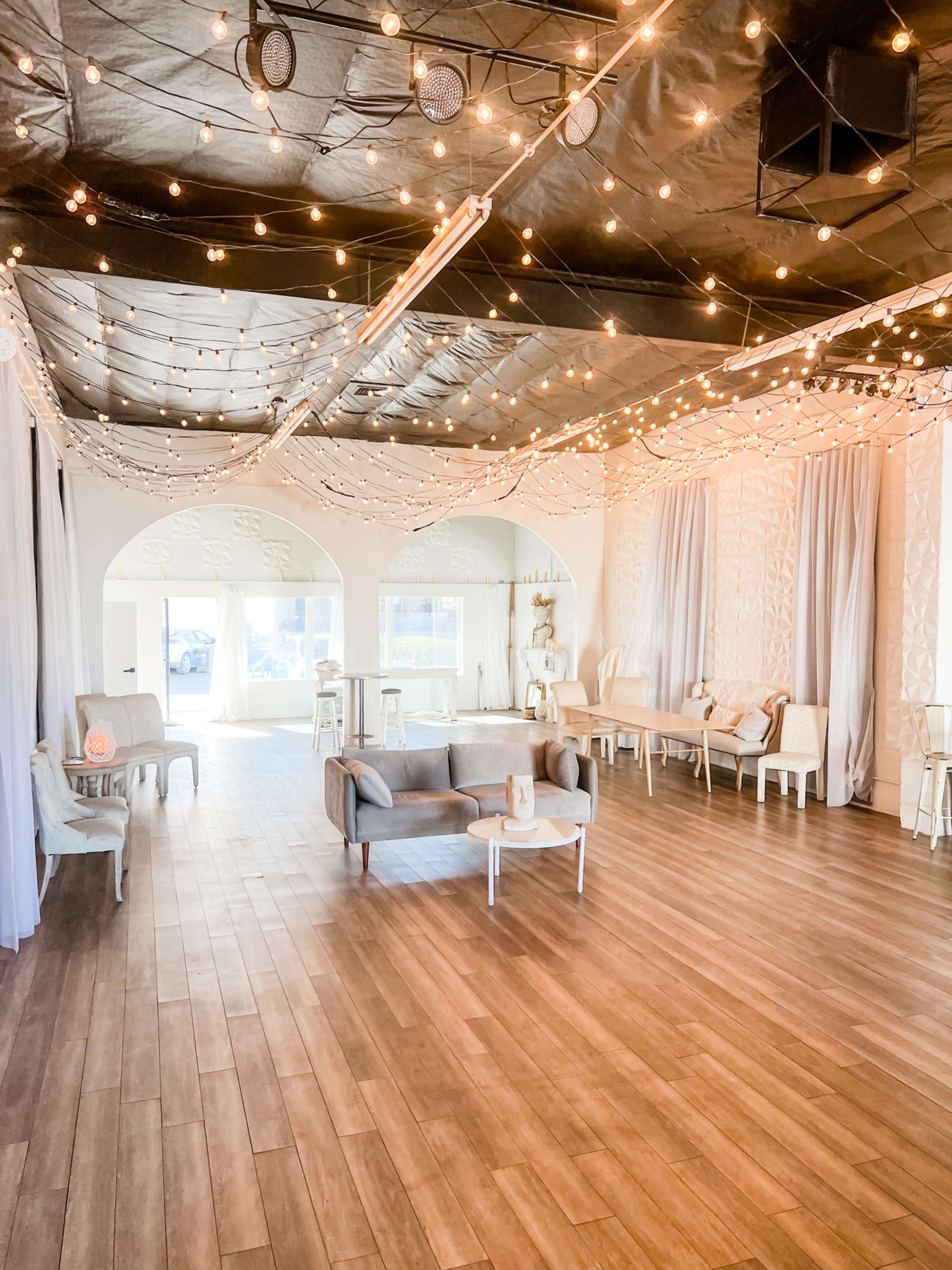 A spacious interior venue adorned with string lights, wooden flooring, and various seating arrangements including a sofa and white chairs.