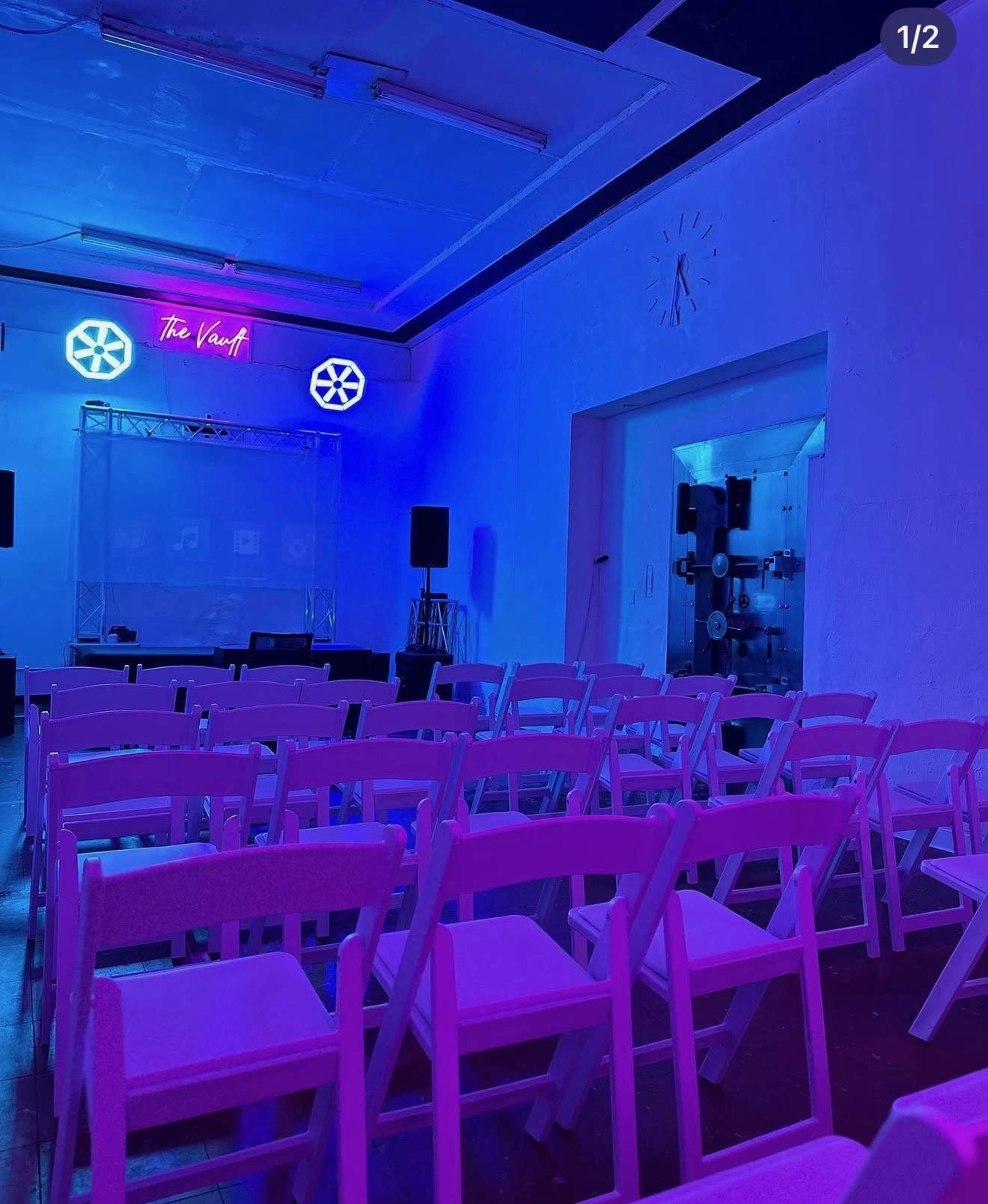 The image shows a dimly lit room with white folding chairs arranged in rows facing a stage, illuminated by blue and pink lighting.