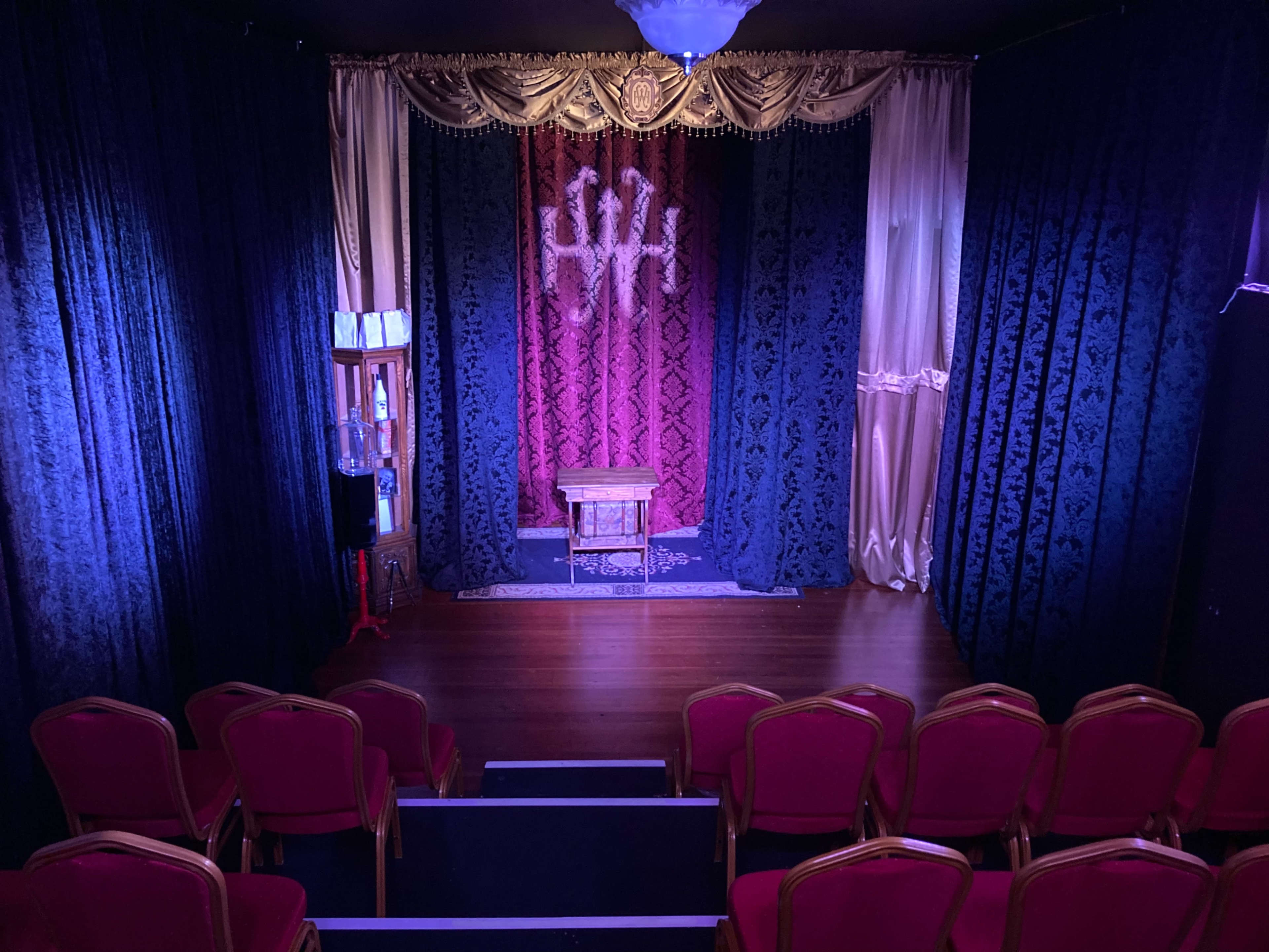 A small theater stage with a decorative backdrop, a wooden table in the center, and red upholstered chairs arranged in front.