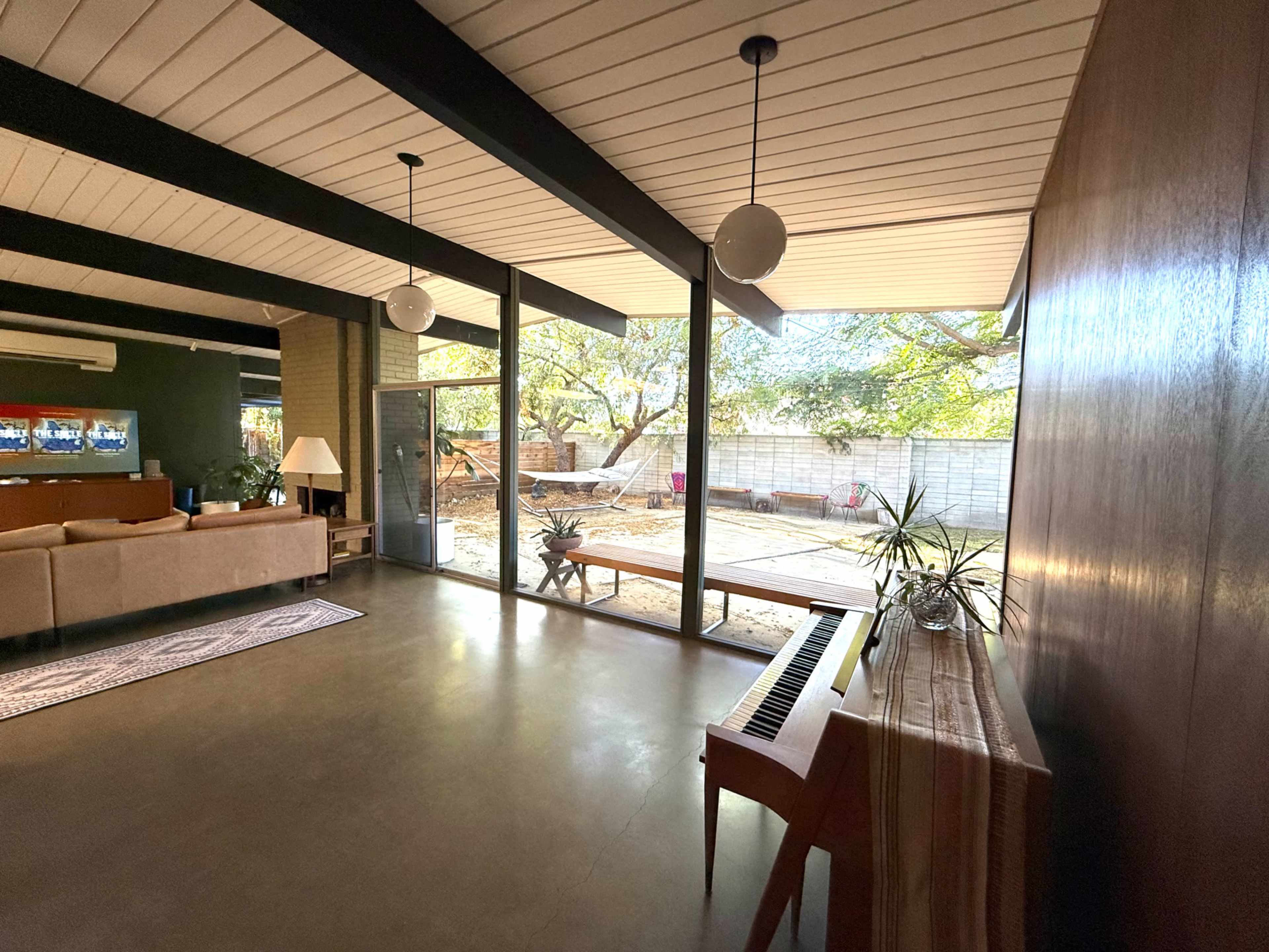 The image shows a living room with a piano, large windows overlooking a yard, and a mix of modern and natural decor.