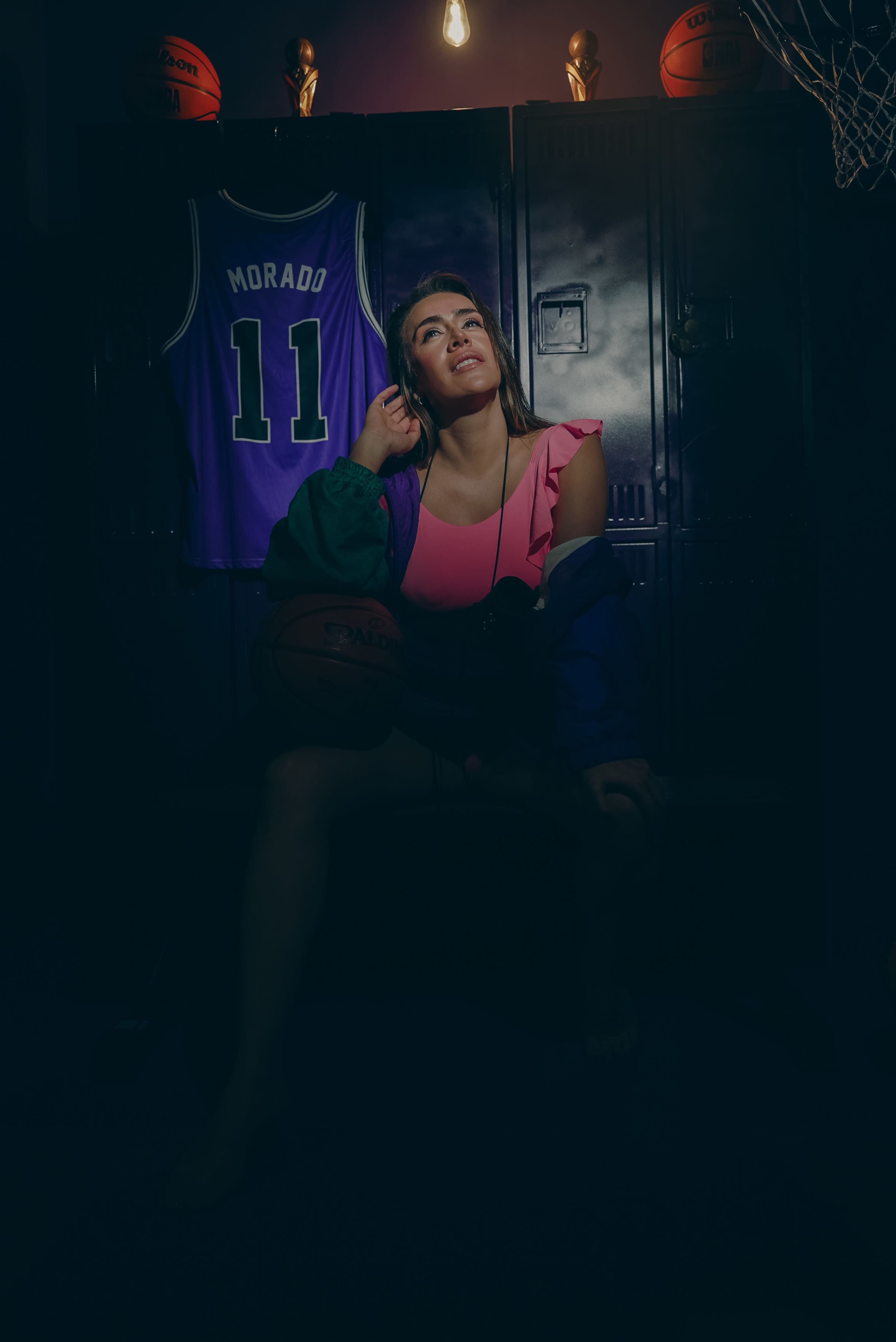 A woman sits on a bench in a dimly lit locker room, holding a basketball, with a purple jersey displayed behind her.