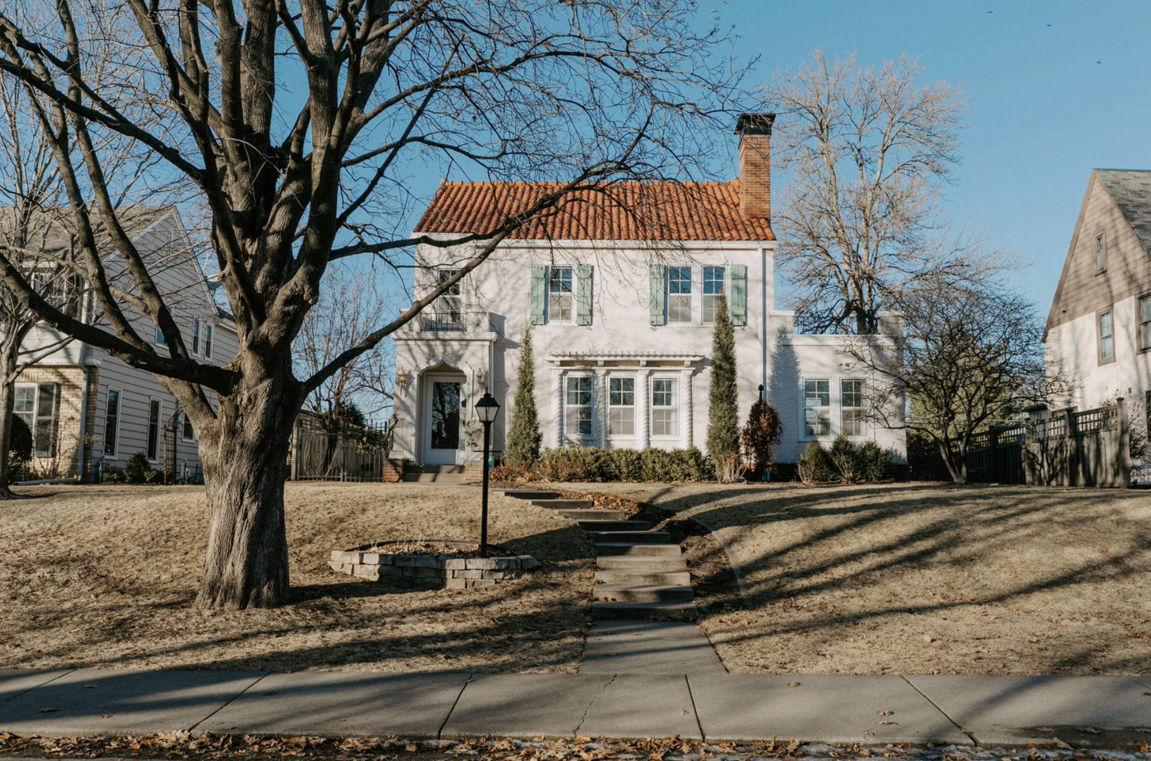 A two-story white house with a red tile roof stands on a grassy lot, flanked by leafless trees and a clear blue sky.