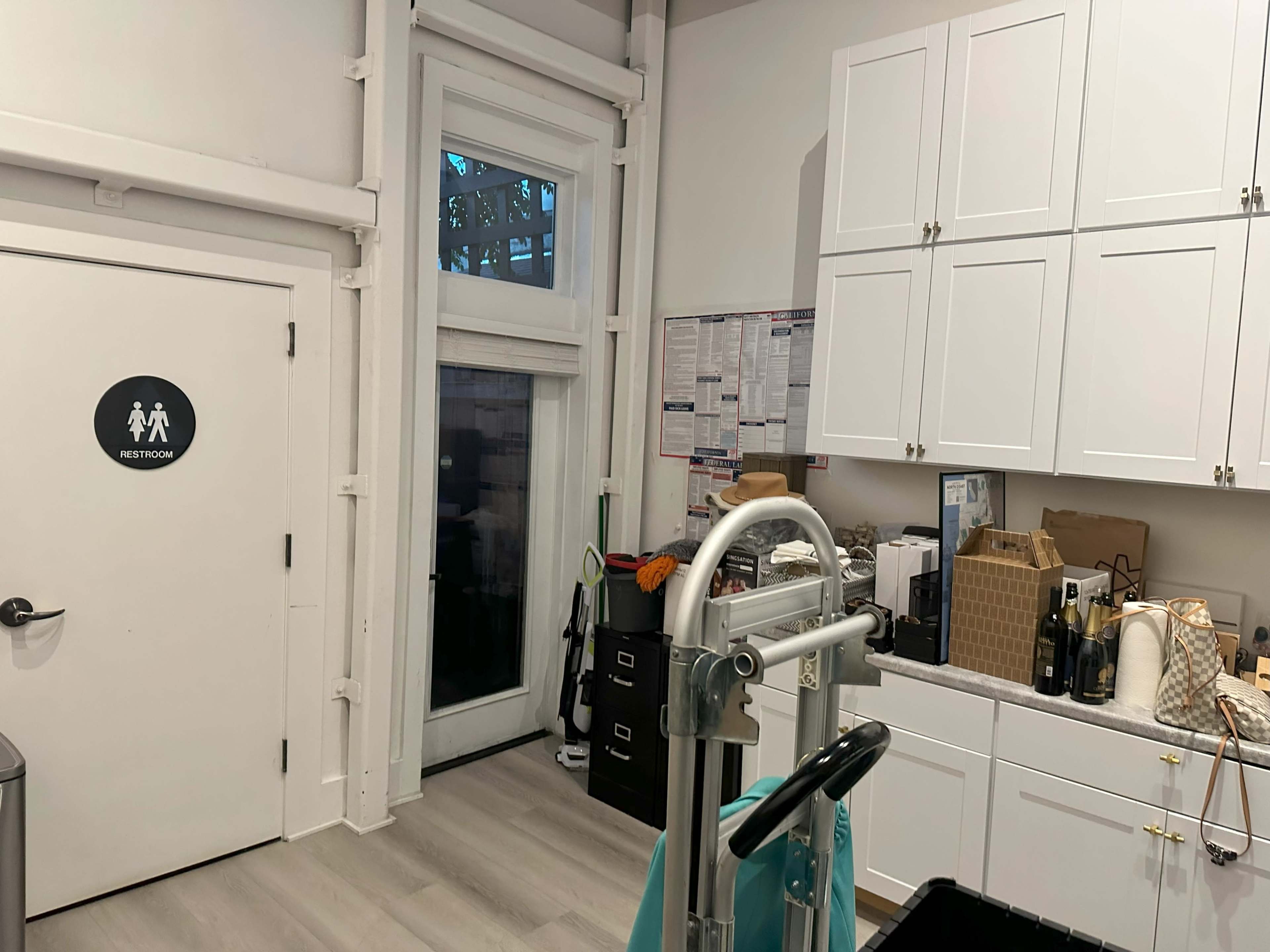 A room with a restroom door on the left, a large window on the right, and various kitchen cabinets and supplies along the wall.
