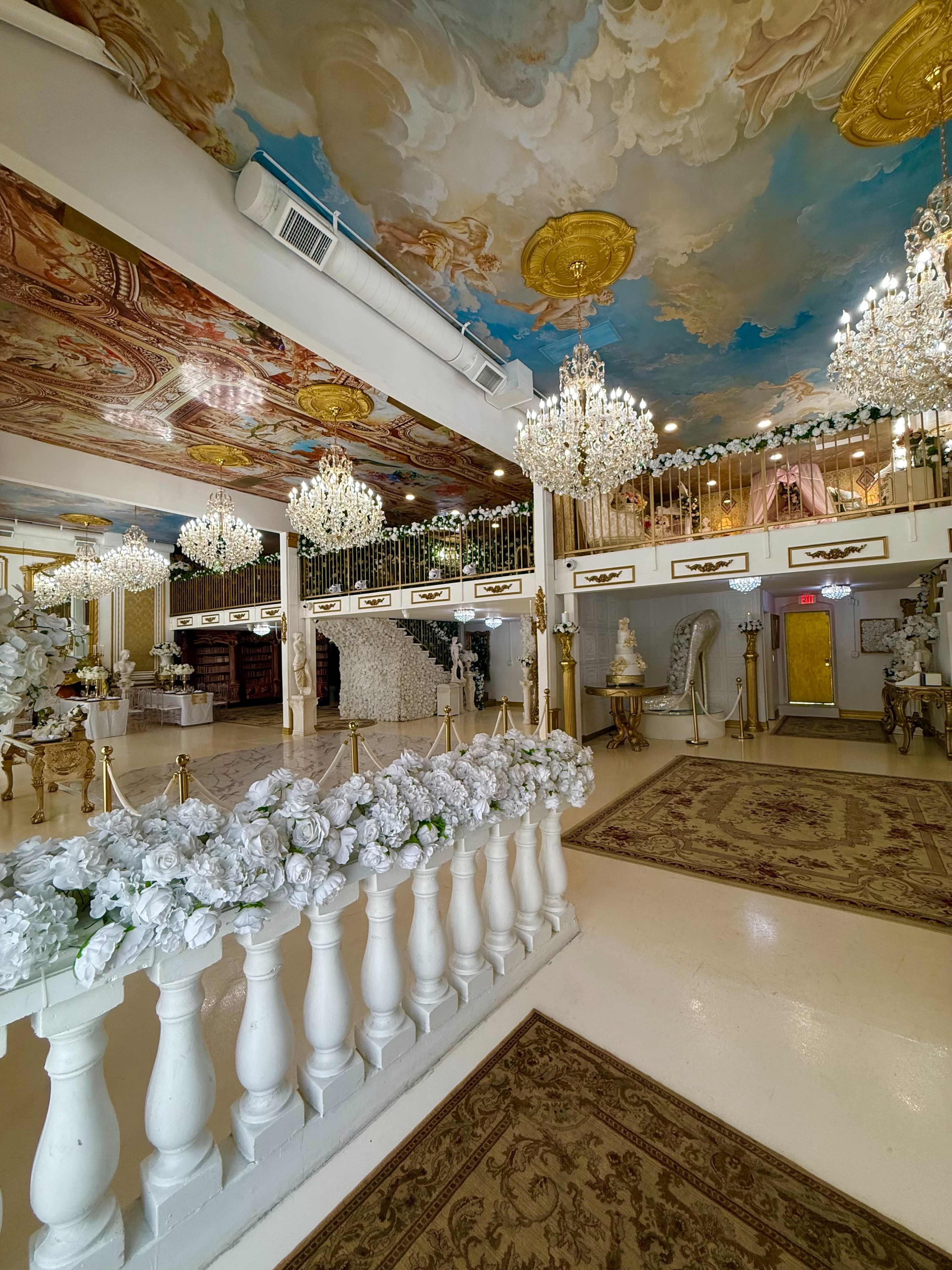 The image shows an ornate interior with a high ceiling adorned with decorative paintings, crystal chandeliers, and floral arrangements along a balustrade.