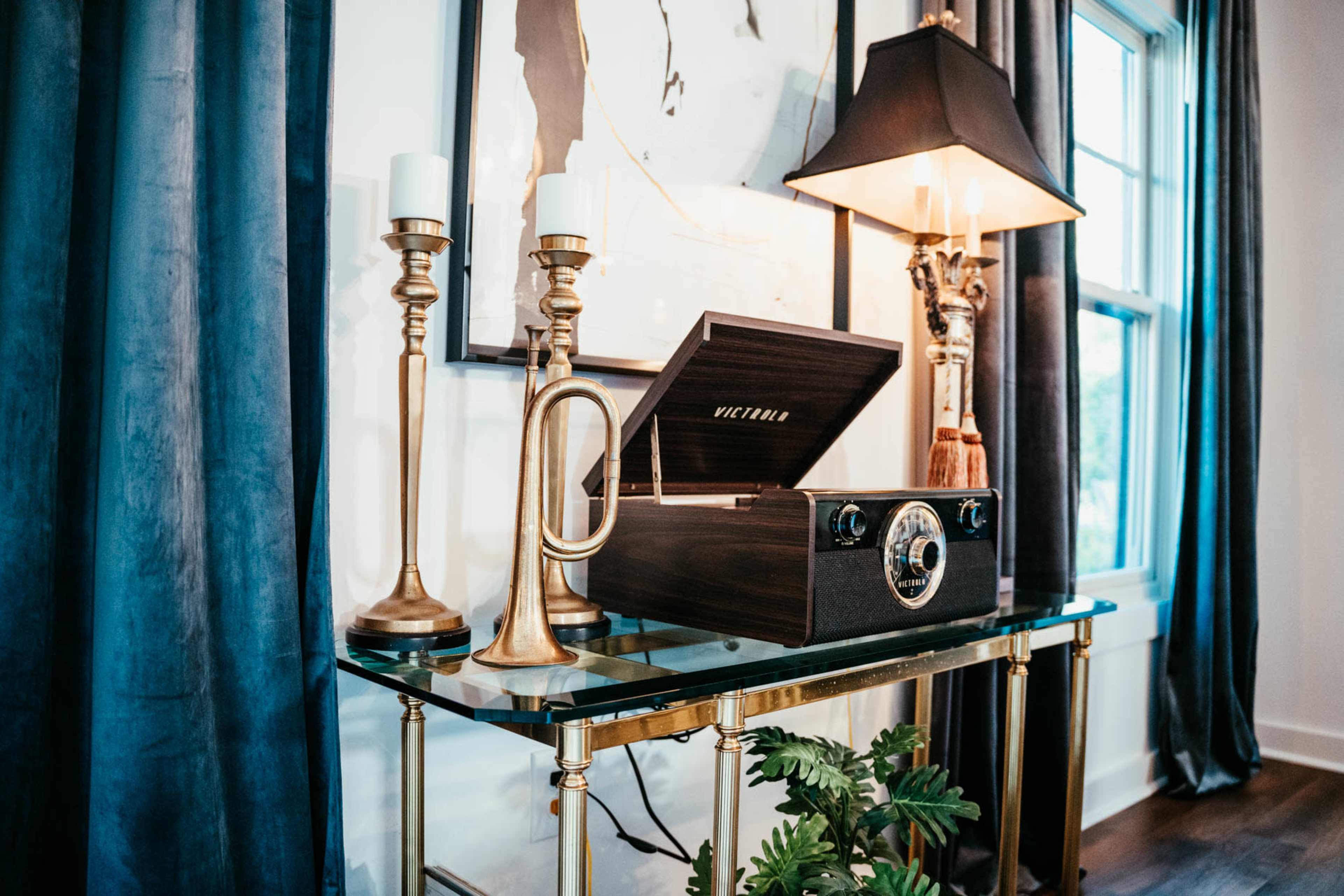 A wooden record player rests on a glass table alongside brass candlesticks and a trumpet, placed near a window with dark curtains.