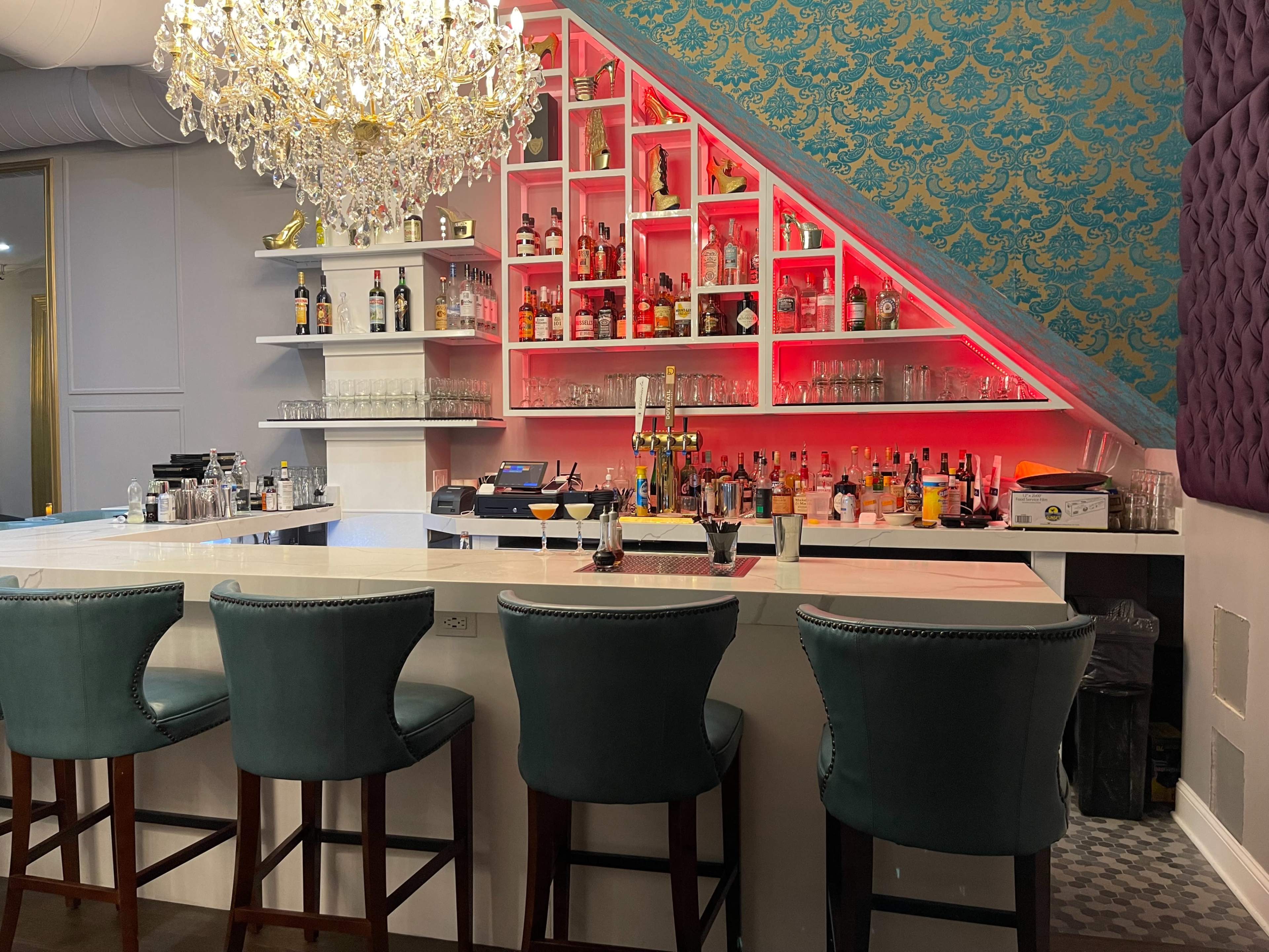 A stylish bar with a decorative chandelier, a backlit shelf displaying various liquor bottles, and upholstered barstools arranged at a marble countertop.