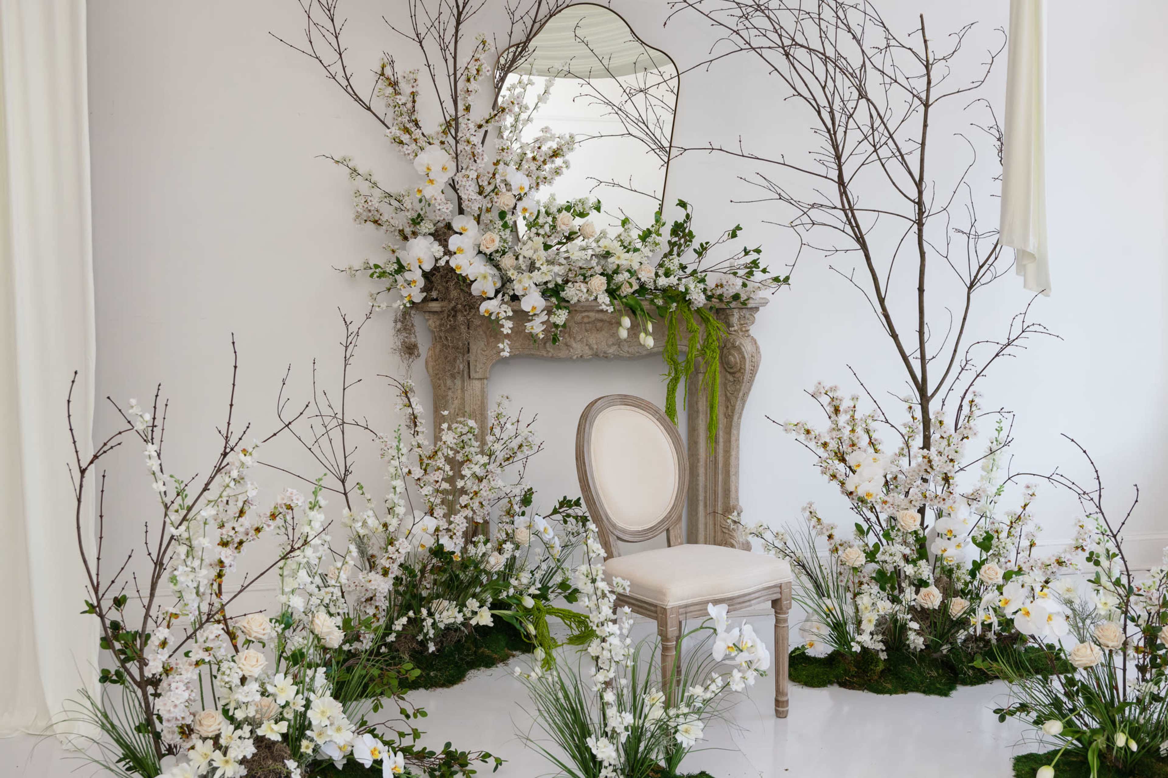 The scene features a decorated room with a vintage mantelpiece, surrounded by white flowering branches and a single elegant chair.