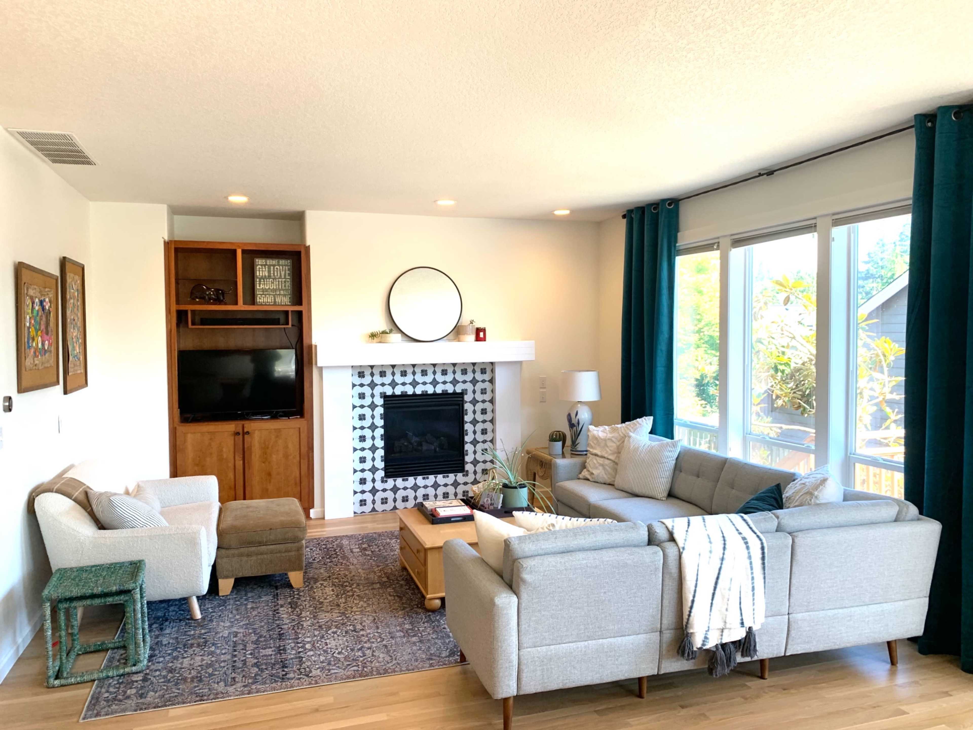 A modern living room features a gray sectional sofa, a wooden coffee table, a patterned area rug, and a fireplace with a circular mirror above it, complemented by large windows and decorative curtains.