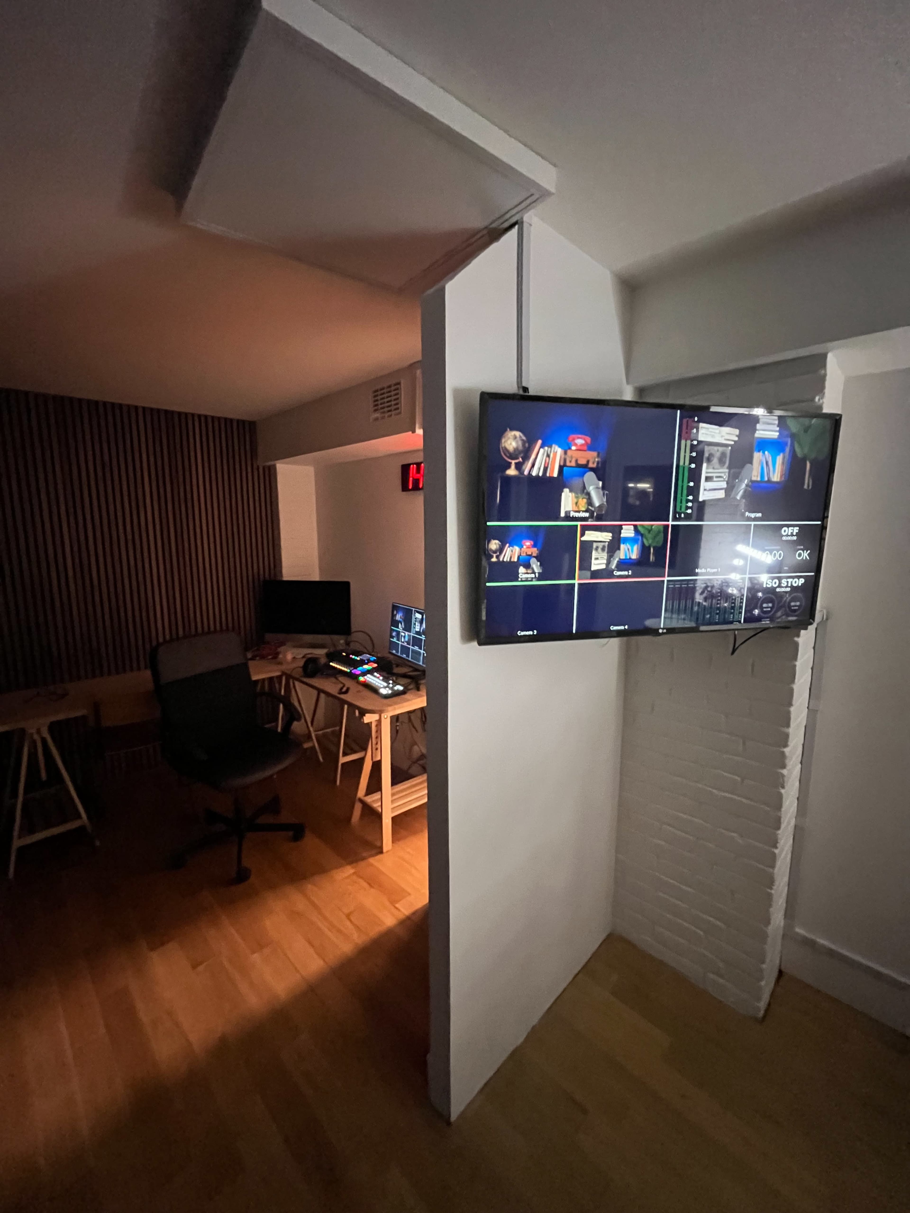 A well-lit room with a large screen displaying various video feeds, adjacent to a desk equipped with computer monitors and seating.