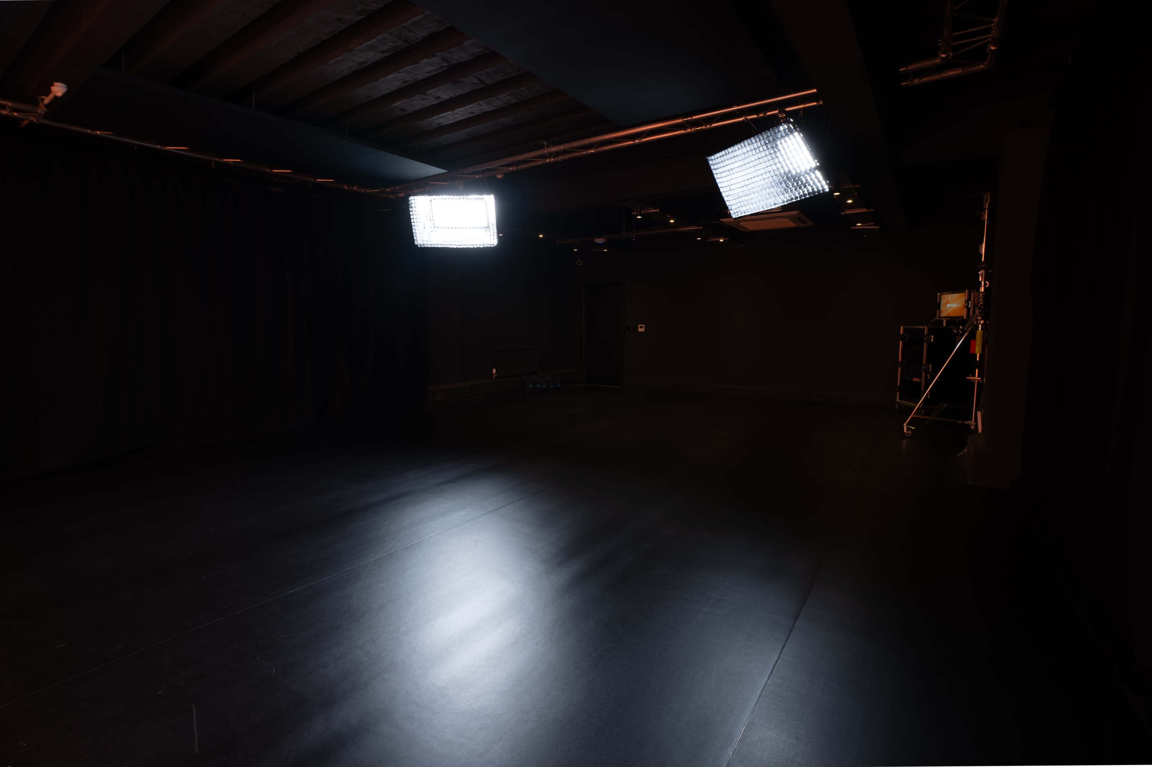 The image shows a dimly lit studio space with black walls and a shiny floor, illuminated by two ceiling-mounted lights.