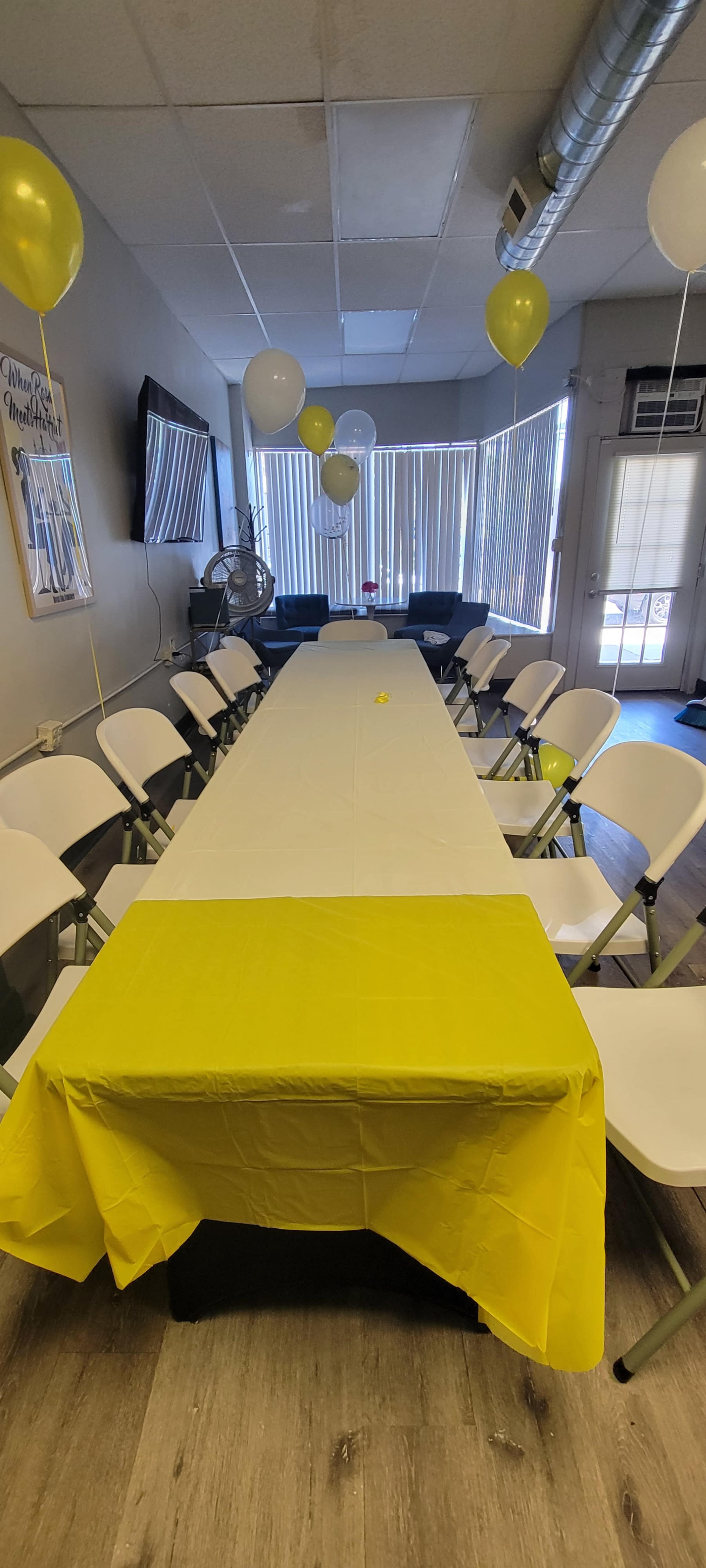 A long table covered with a yellow tablecloth is set up in a room with white chairs and decorated with balloons.