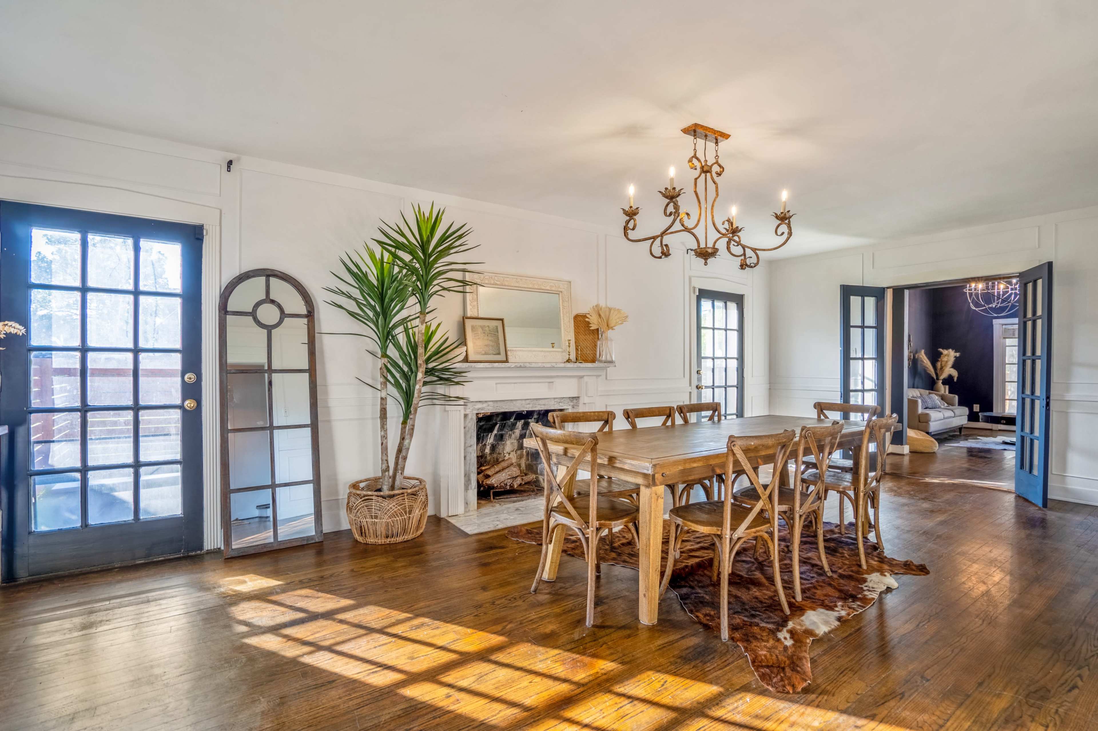 A spacious dining room with a wooden table surrounded by chairs, a chandelier overhead, and large windows allowing natural light to enter, alongside a fireplace and decorative elements.