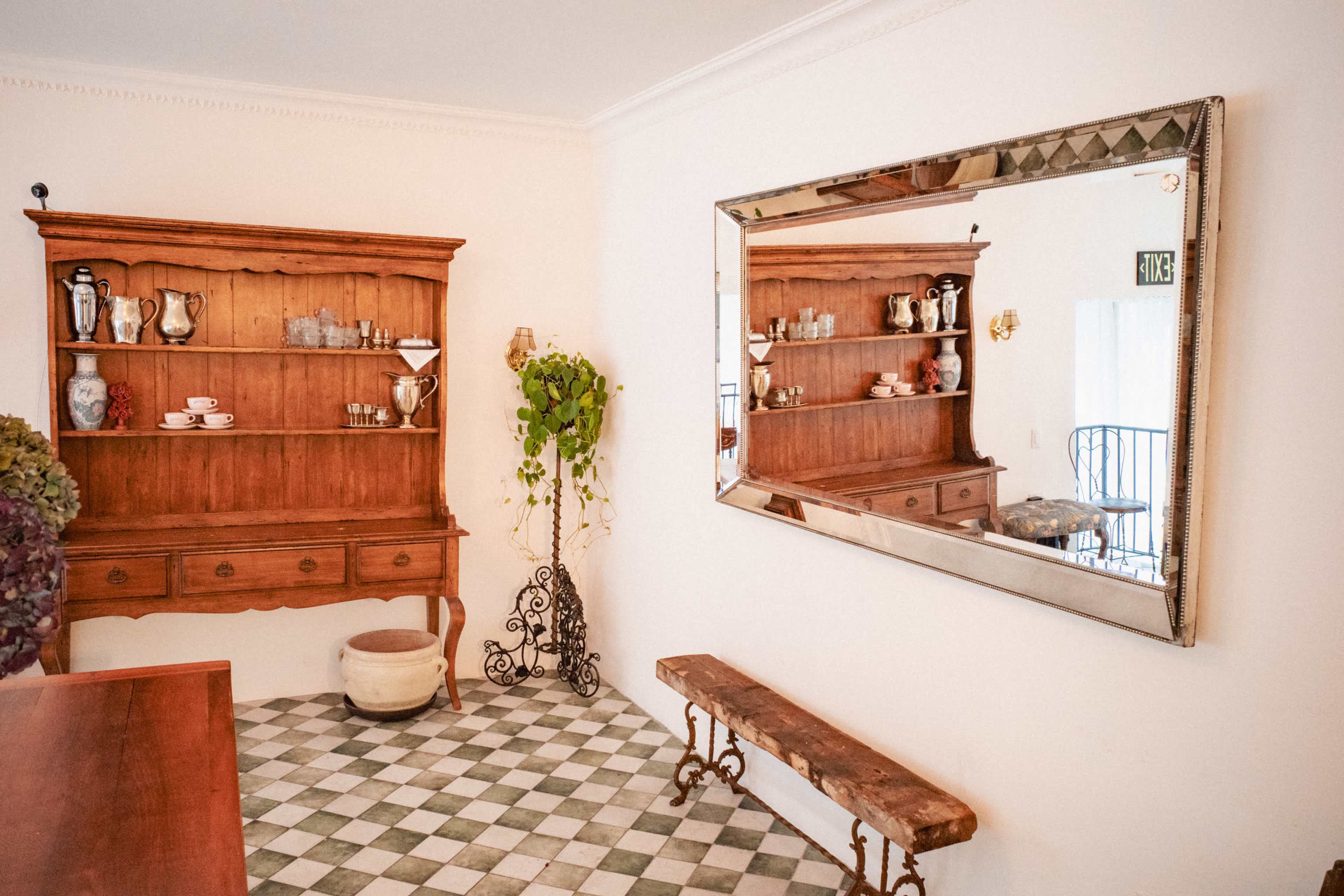 The image shows a room featuring a wooden hutch filled with dishware, a large mirror mounted on the wall, a stone planter, and a wooden bench on a checkered floor.