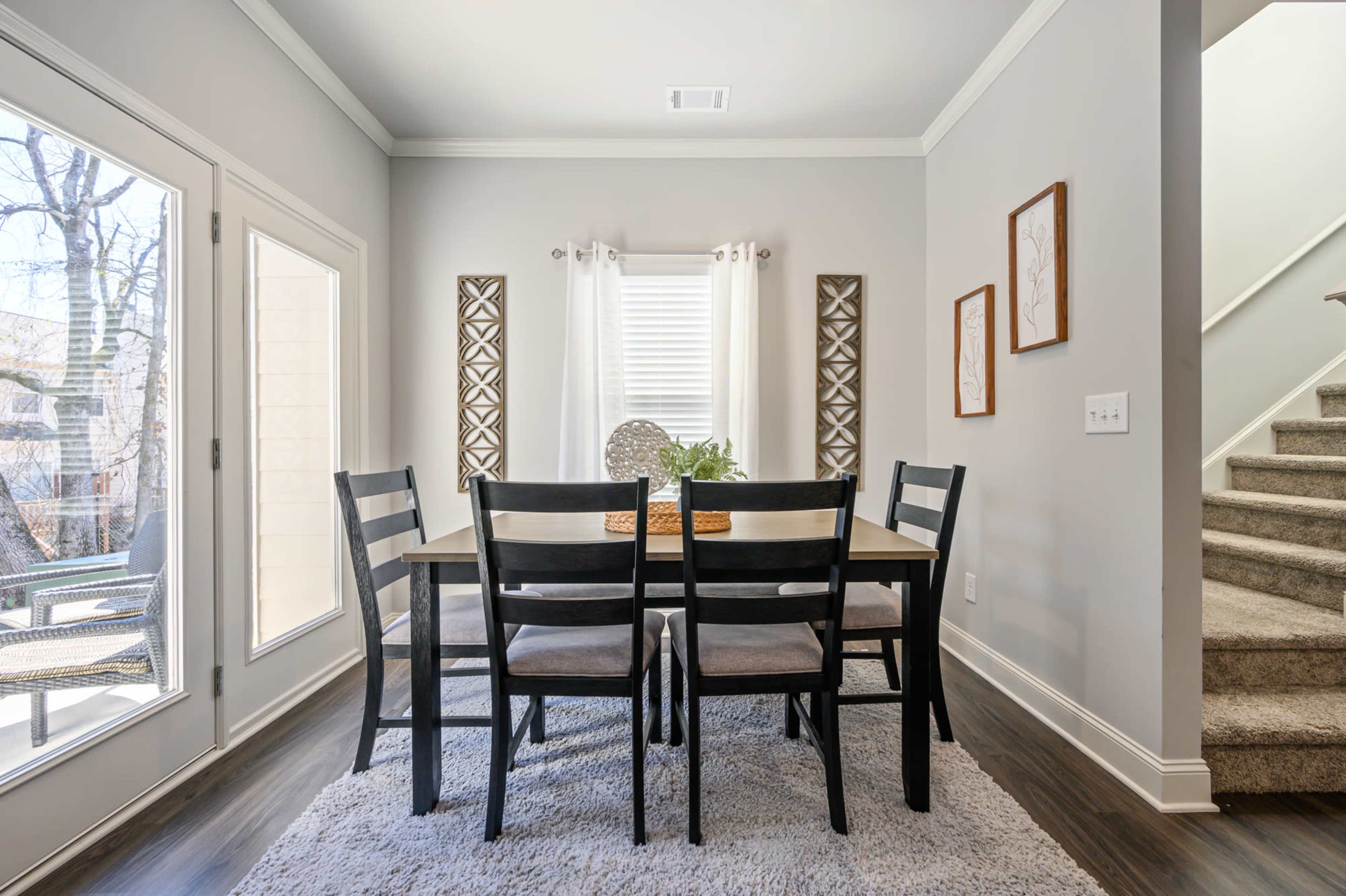 A dining area features a rectangular table surrounded by four chairs, with large windows and a staircase in the background.