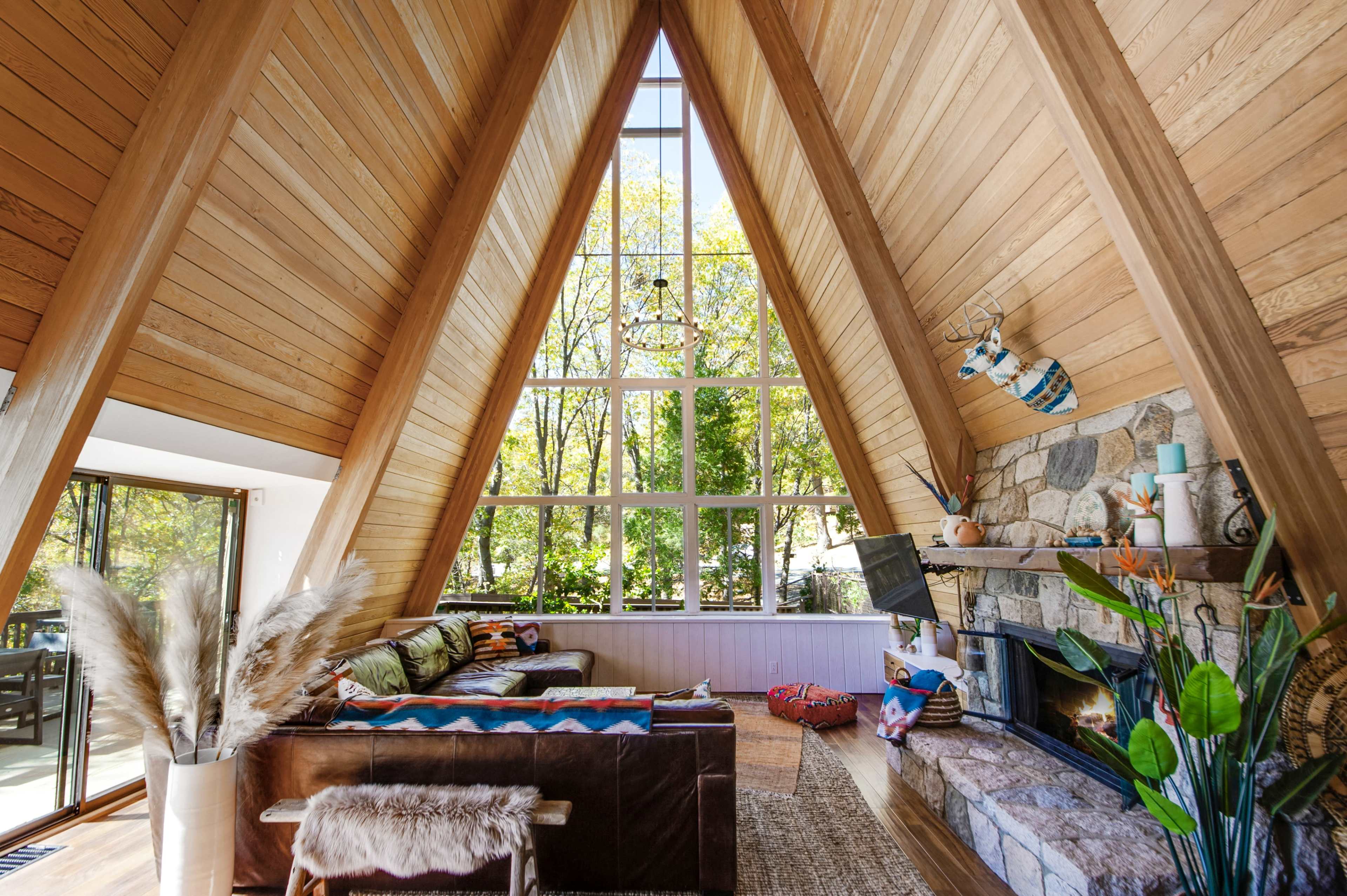The interior of an A-frame cabin features tall wooden ceilings, large windows, and a stone fireplace surrounded by comfortable seating.