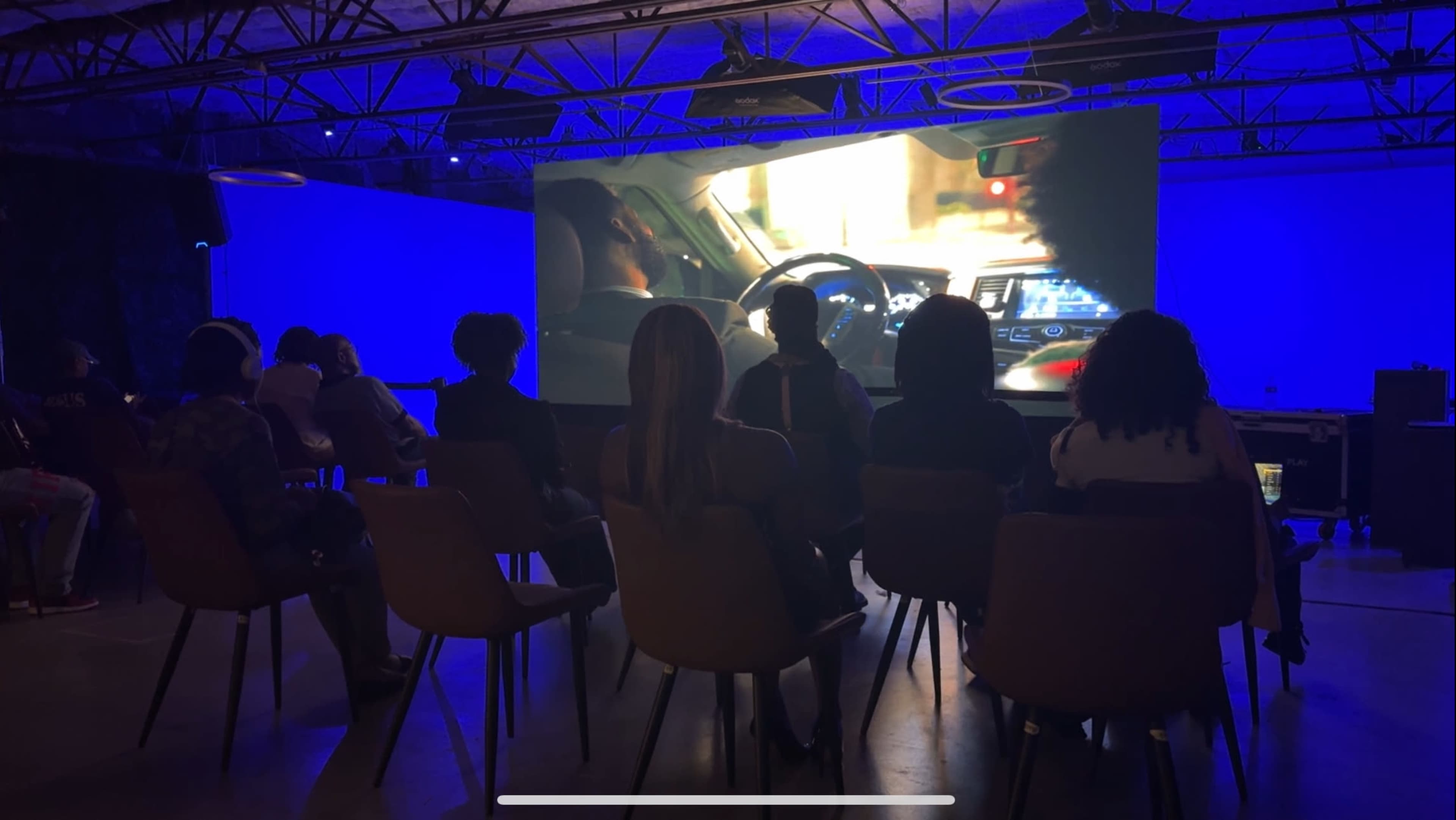 A group of people sits in chairs, facing a large screen that displays a scene from a movie or video, in a dimly lit room with blue ambient lighting.