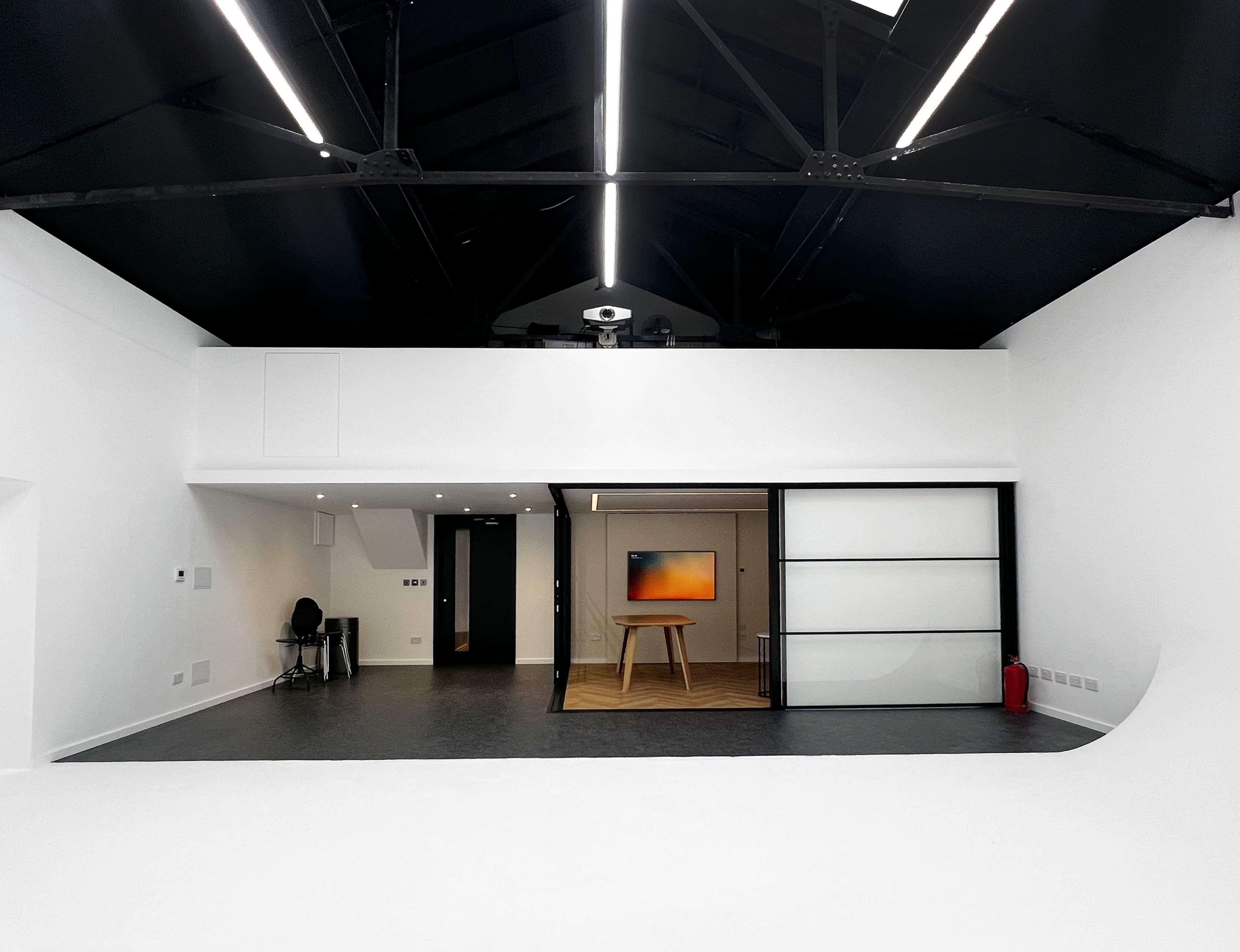 The image shows a modern, open space with high ceilings featuring black beams, a polished floor, and a glass door leading to a room with a table and abstract artwork.