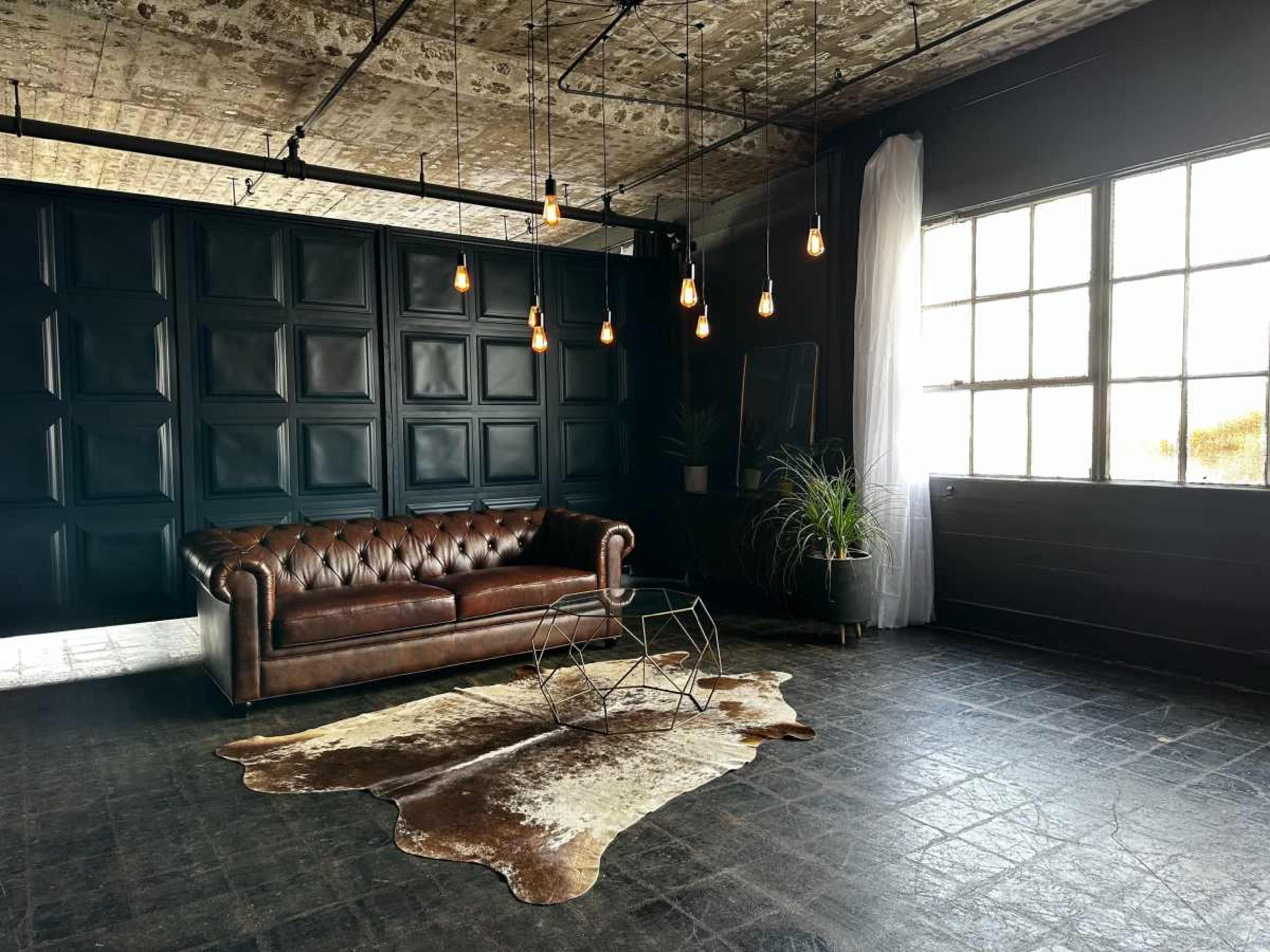 The image shows a modern interior featuring a brown leather chesterfield sofa, a cowhide rug, and hanging light fixtures against a dark paneled wall.