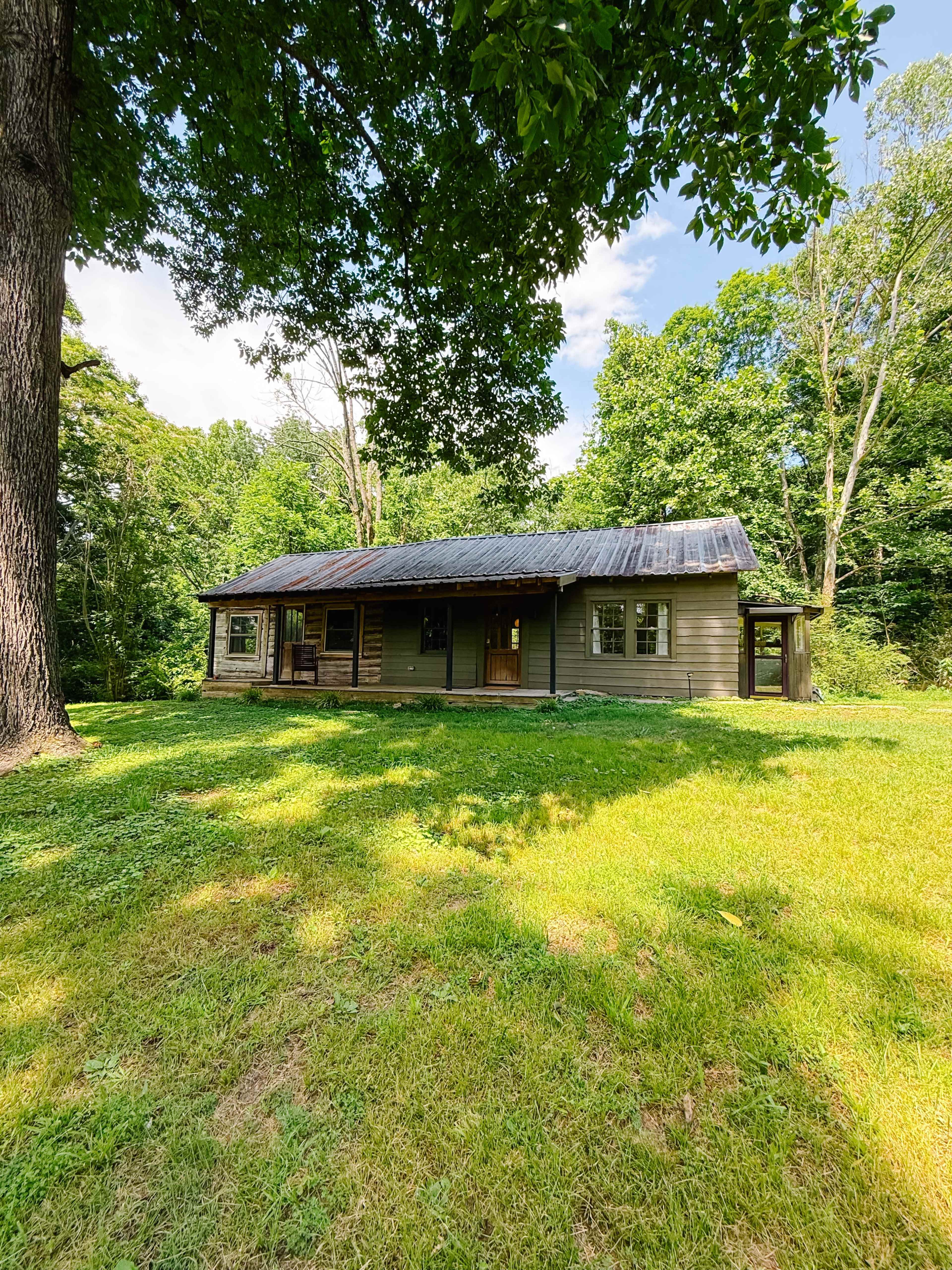 The image shows a single-story cabin with a metal roof, surrounded by green grass and trees.