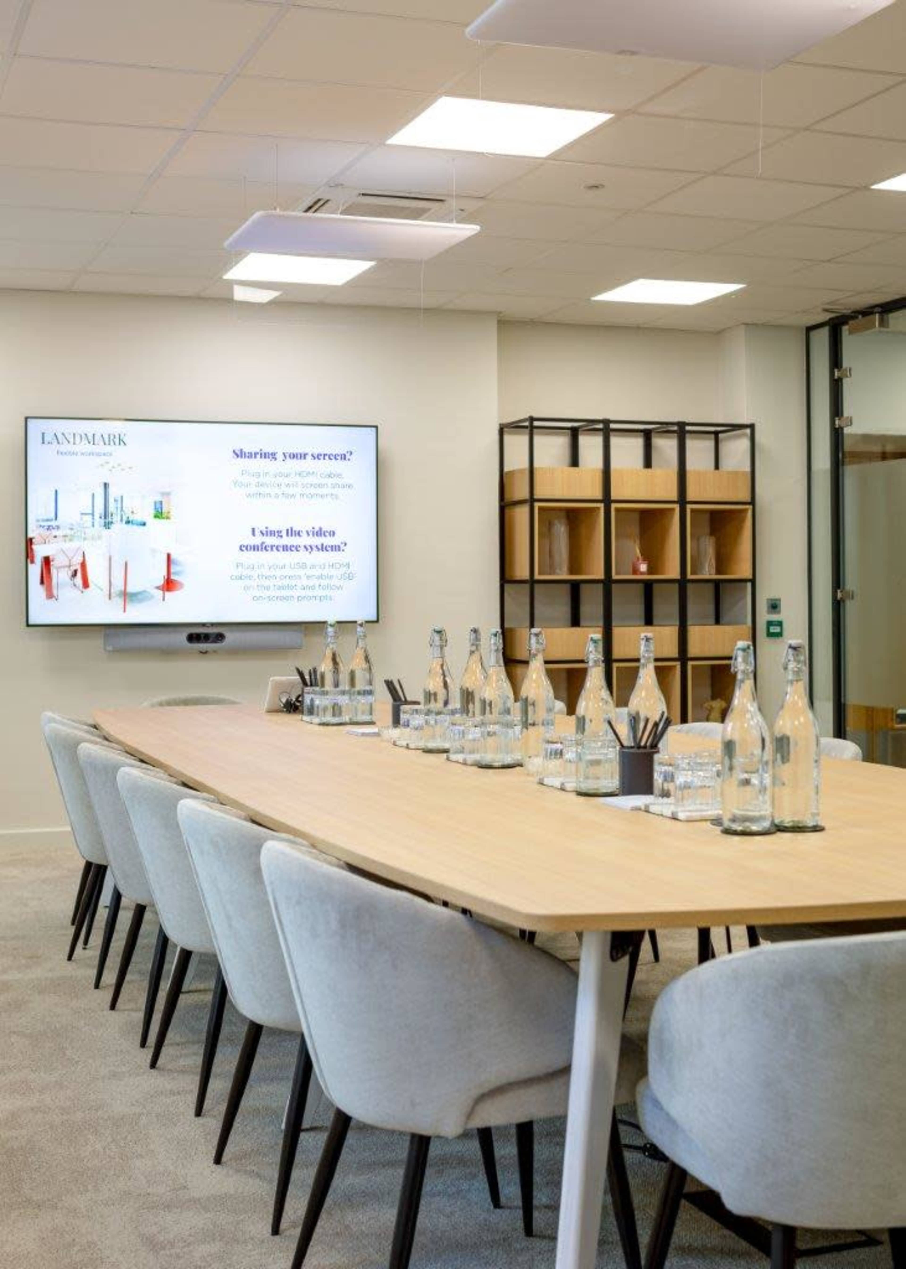 The image shows a modern conference room with a long wooden table, styled chairs, a presentation screen, and several glass water bottles arranged neatly on the table.