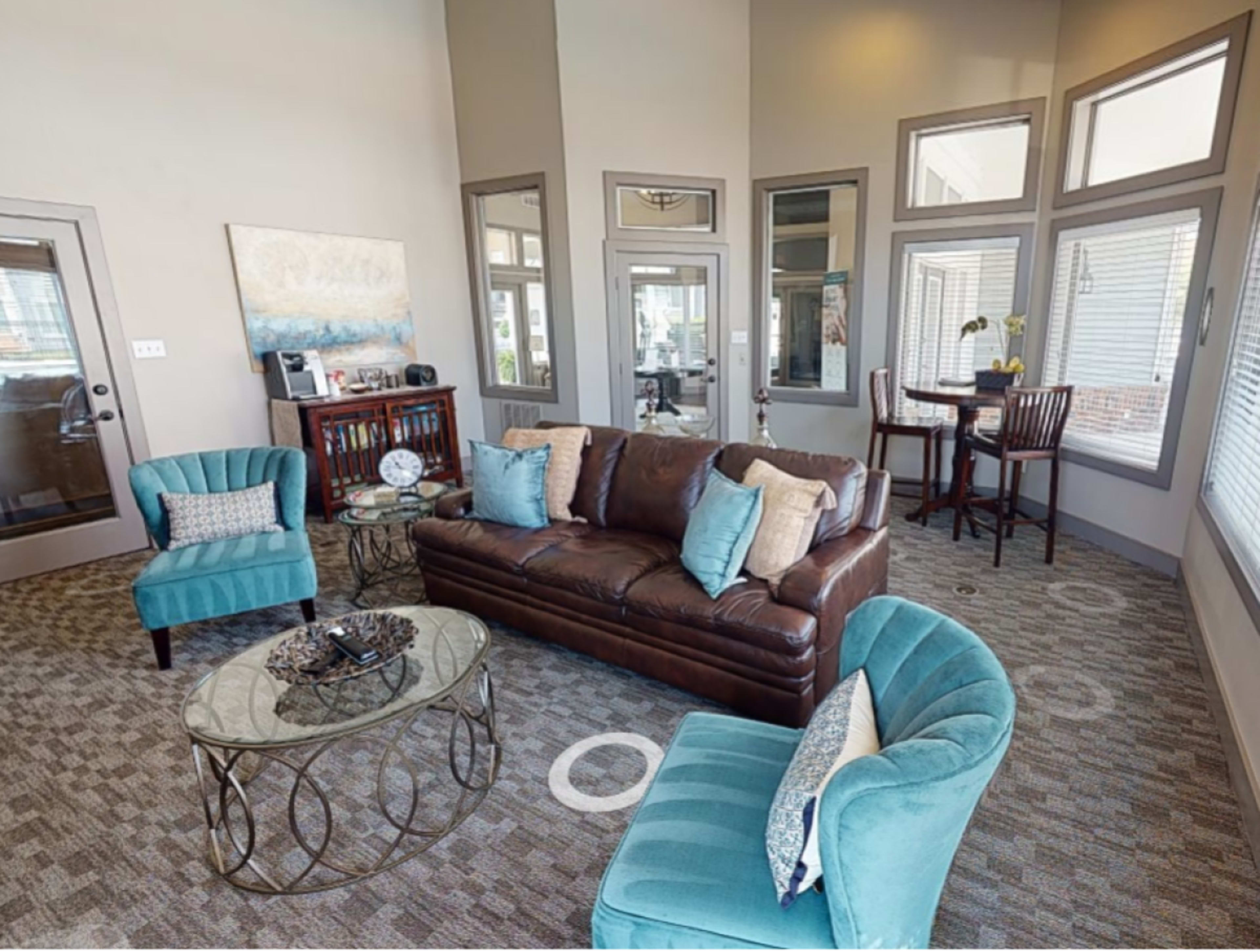 A modern lounge area featuring a brown leather sofa, turquoise armchairs, a glass coffee table, and a small dining table with chairs.