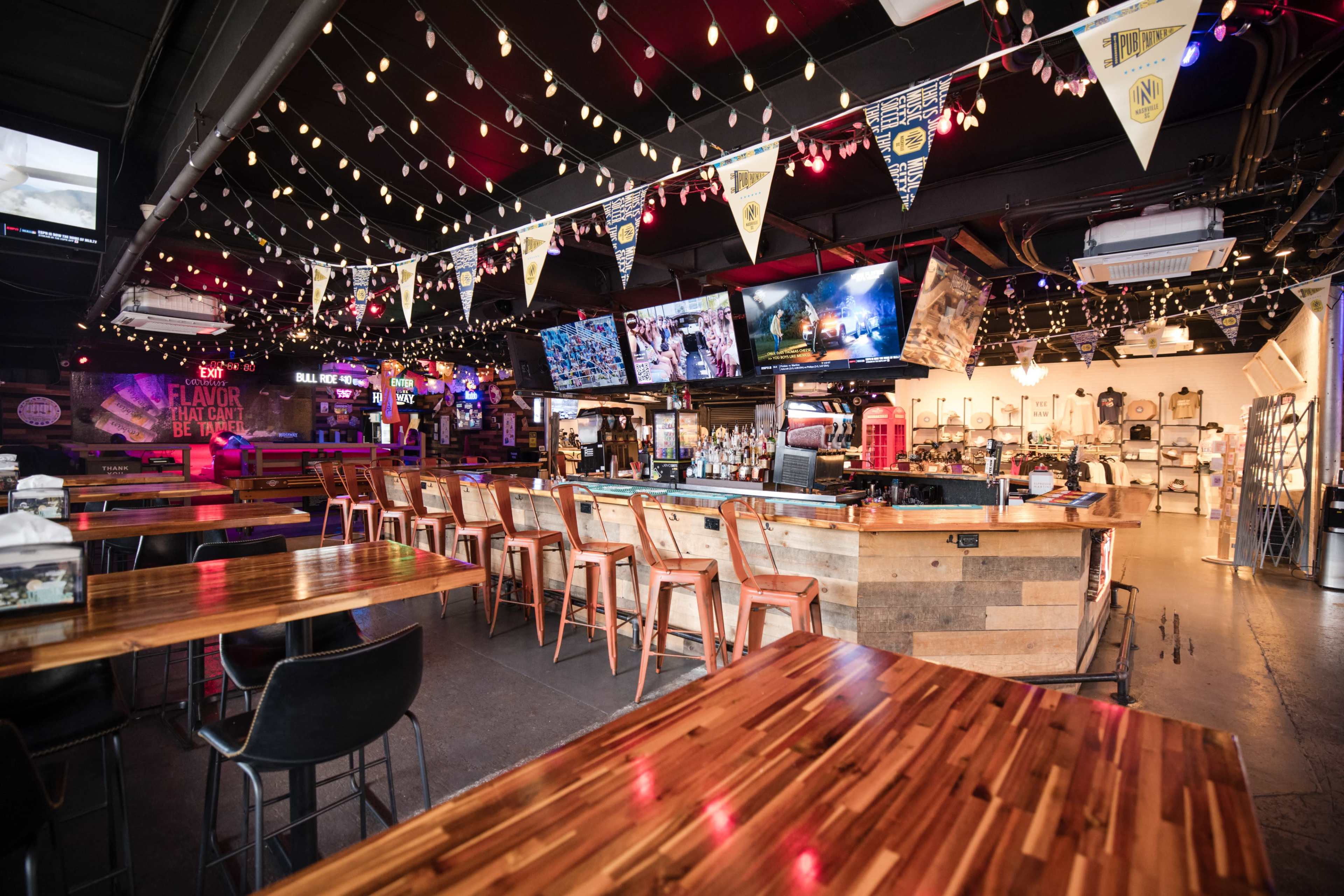 A modern bar and dining area features a wooden countertop, high stools, multiple televisions displaying sports, and decorative lights overhead.