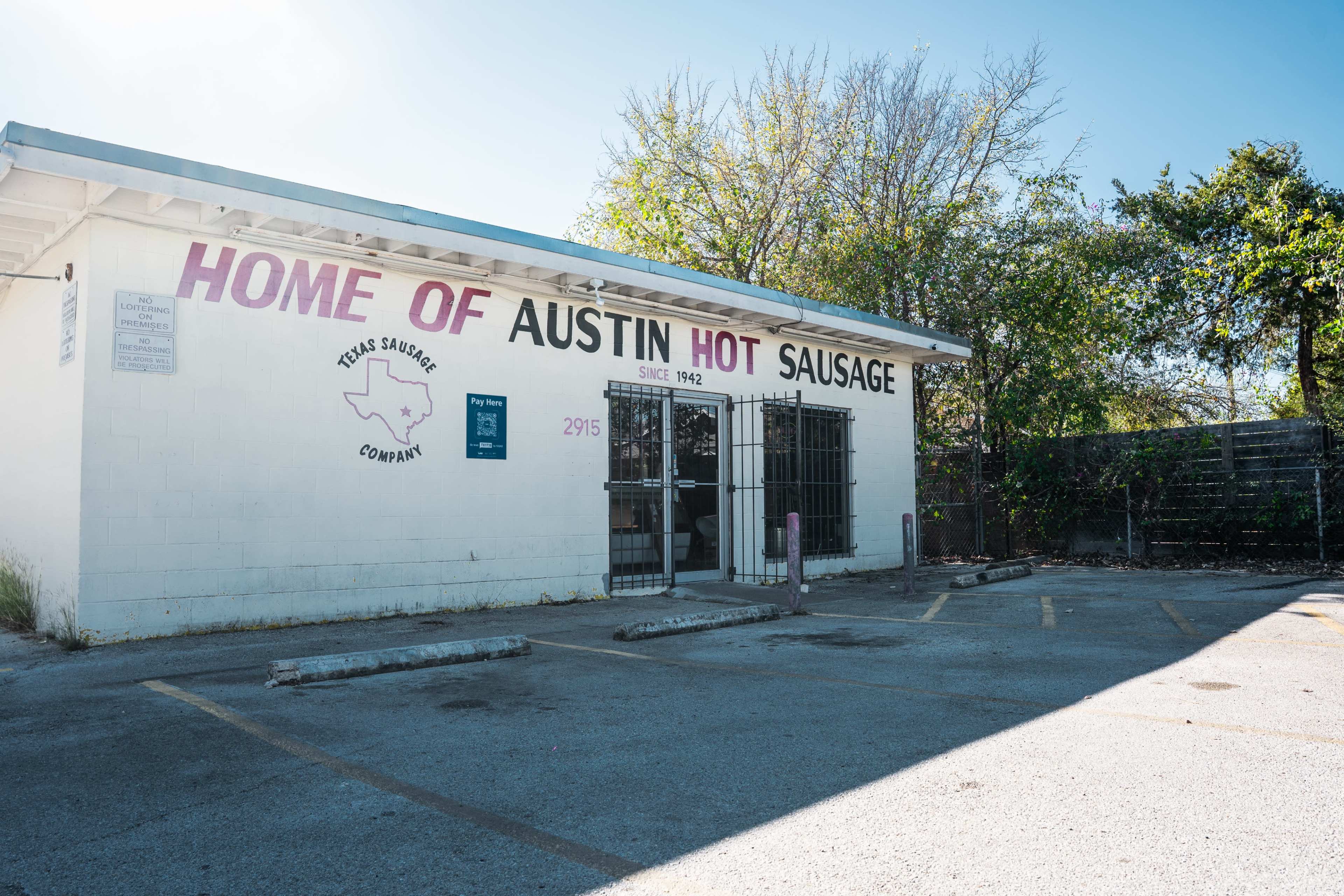 The building displays a sign that reads "Home of Austin Hot Sausage" and features a simple, white exterior with a parking lot in front.