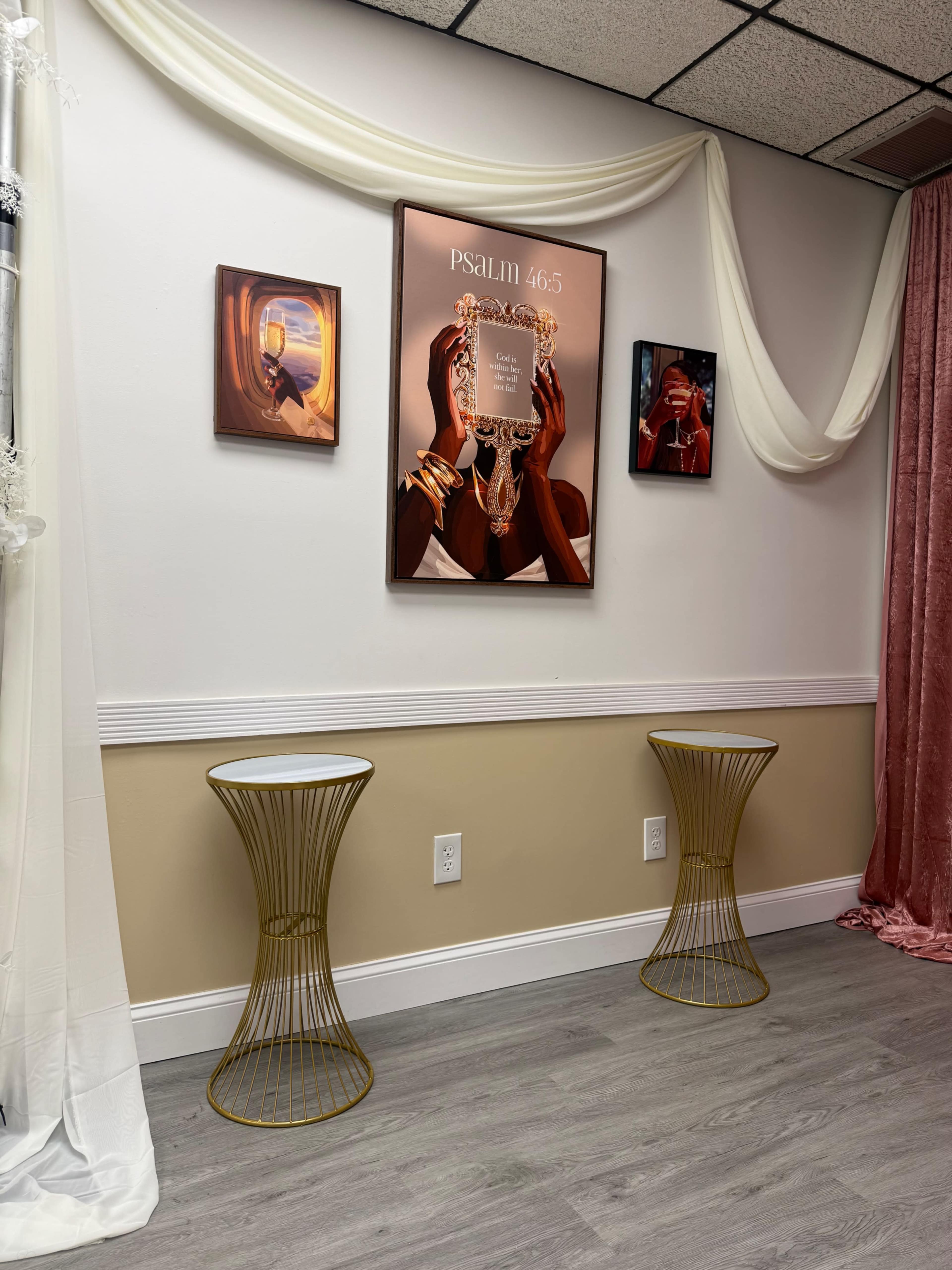 The image features a wall adorned with three pieces of artwork, including a prominent framed piece of art, and two gold wire side tables positioned in front on a light wood floor.