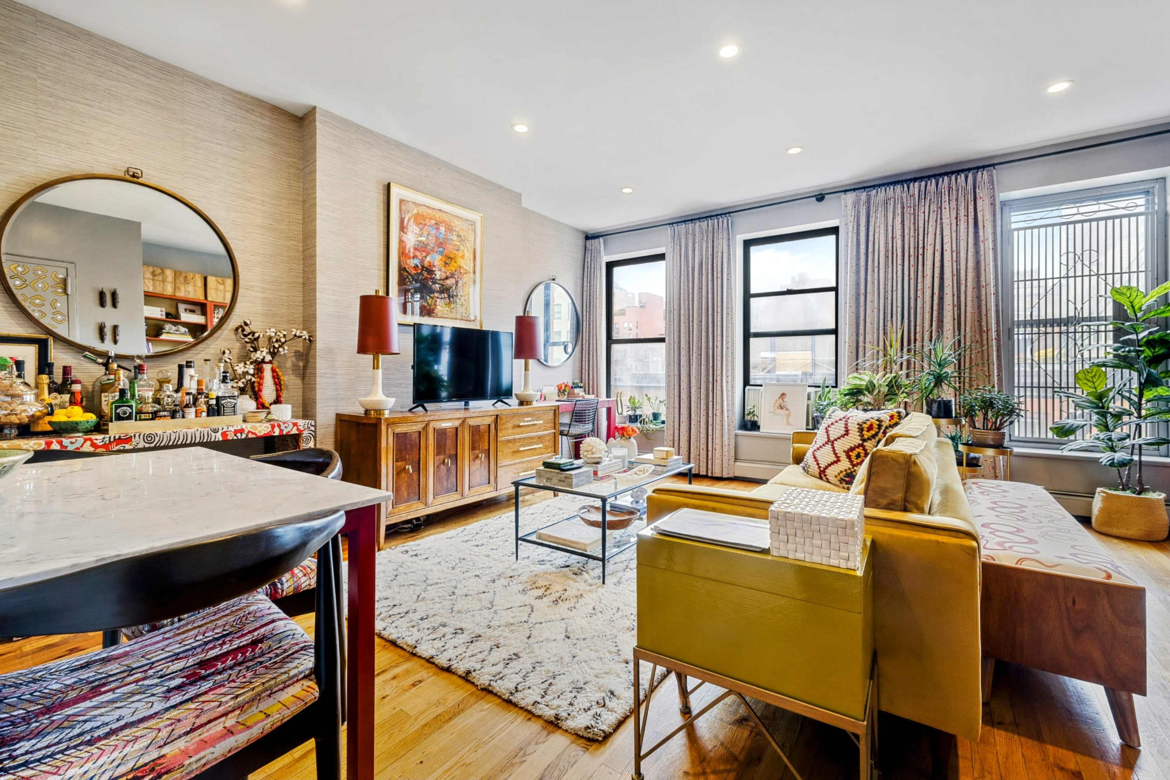 A cozy living room features a mix of plants, a television, and a vintage wooden sideboard, with large windows allowing natural light to fill the space.