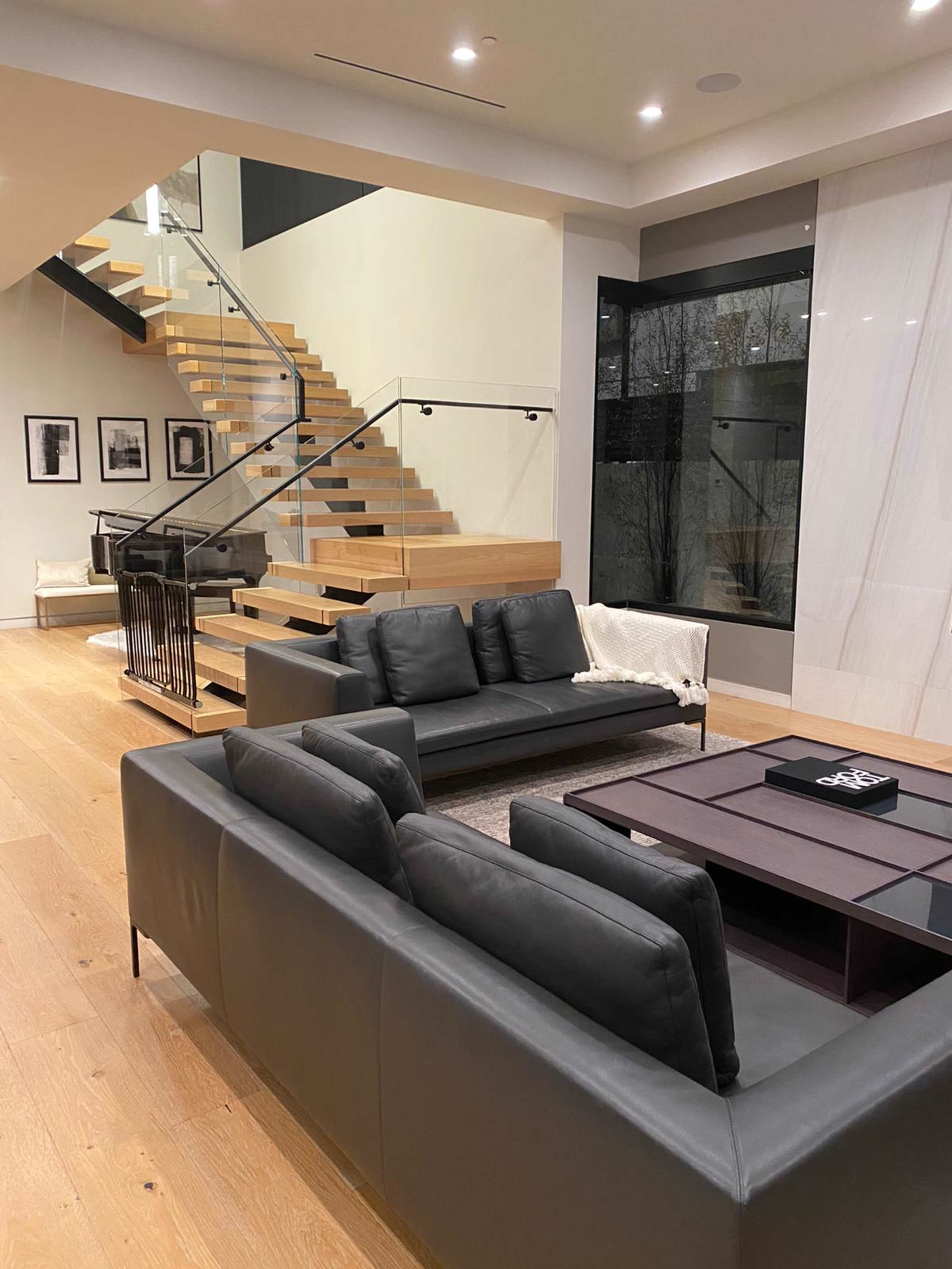 The image shows a modern living room with a dark leather sofa, a coffee table, and a wooden staircase leading to an upper level.