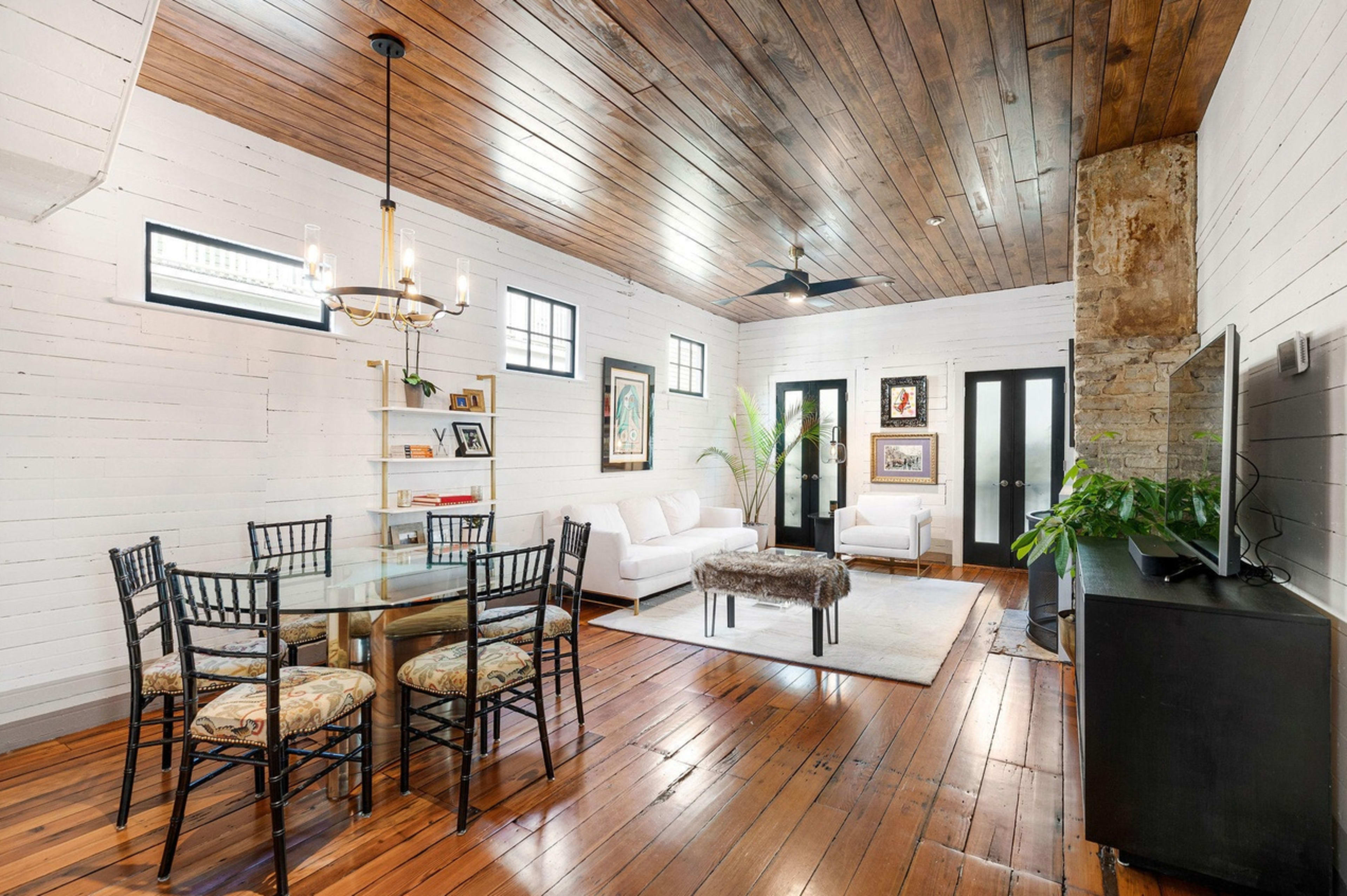 A modern living space featuring a dining area with a glass table and black chairs, a white sofa, and wooden flooring with a high ceiling.