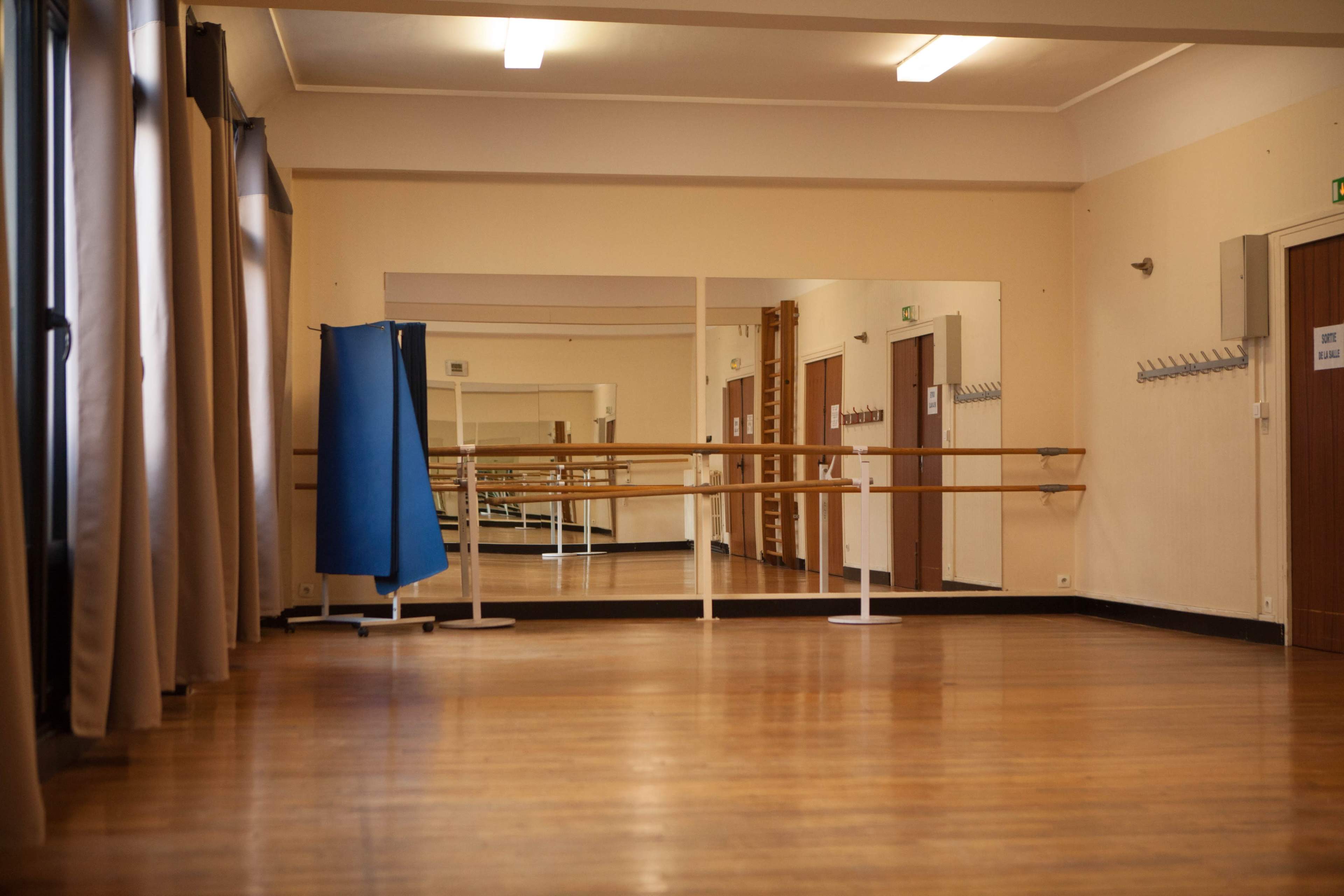 A spacious dance studio features wooden flooring, mirrors along one wall, and a blue partition in the corner.