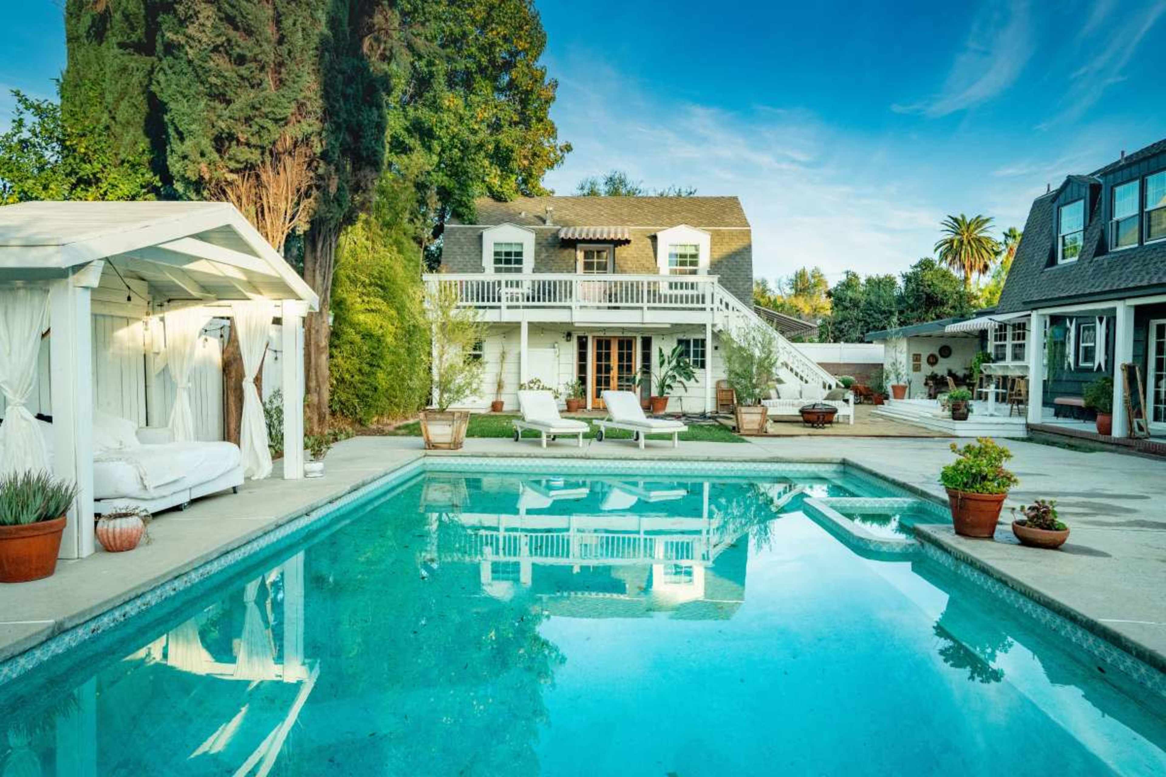 The image shows a serene backyard with a swimming pool, surrounded by lush greenery and two houses in the background, one featuring a balcony.