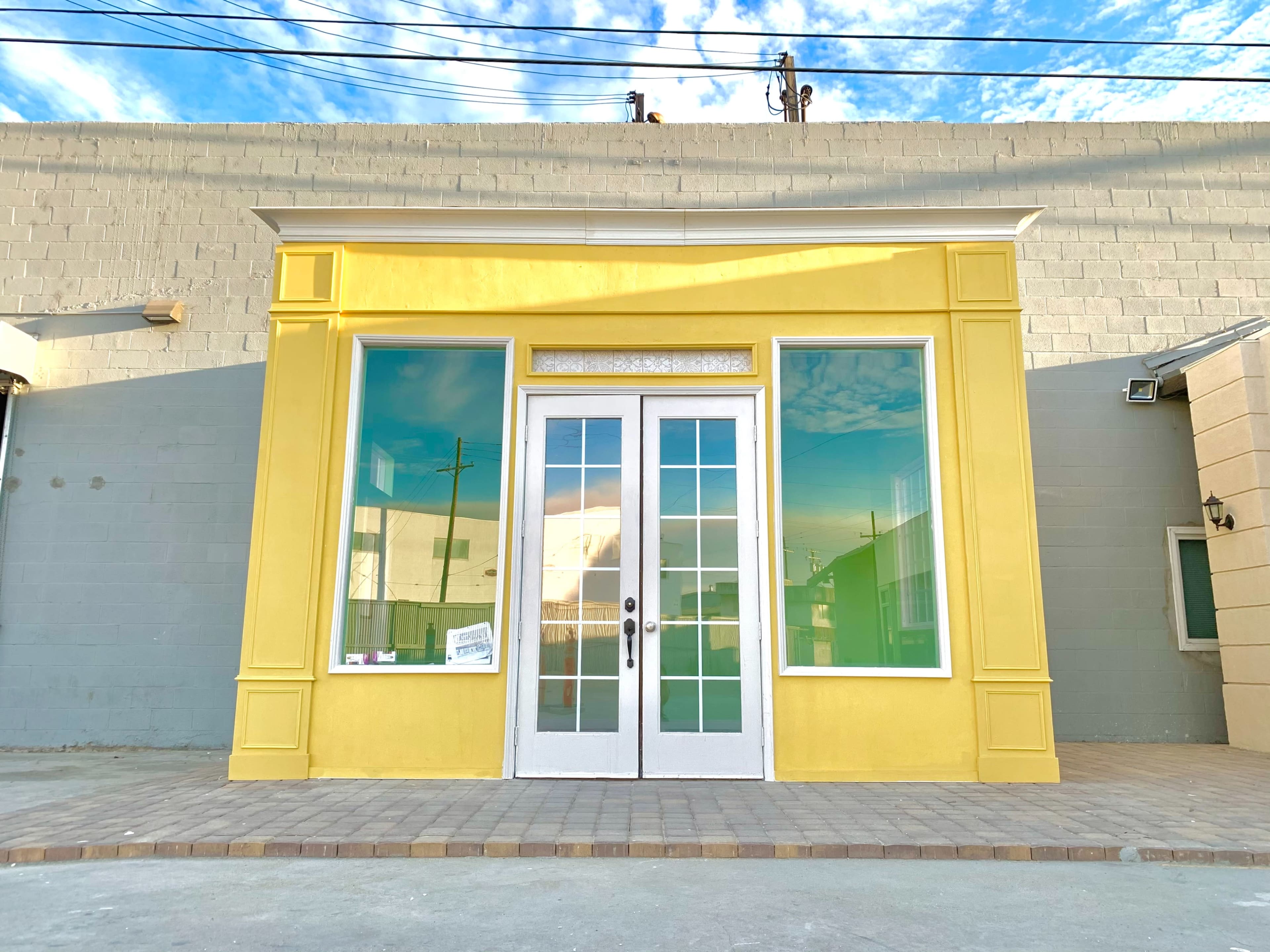 A bright yellow storefront with large windows and double doors reflects the sky and surrounding buildings.