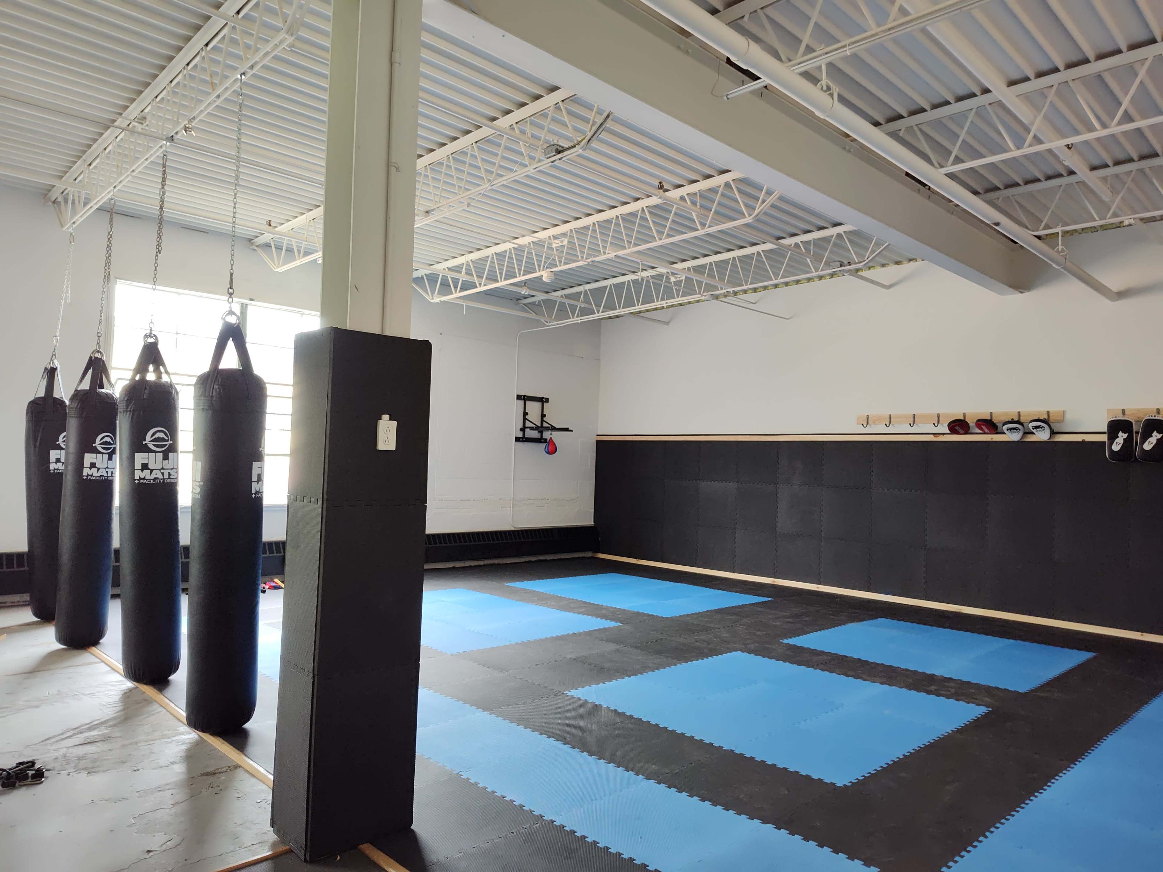 The image shows a spacious martial arts studio with hanging punching bags, black and blue mats, and a wall-mounted training apparatus.