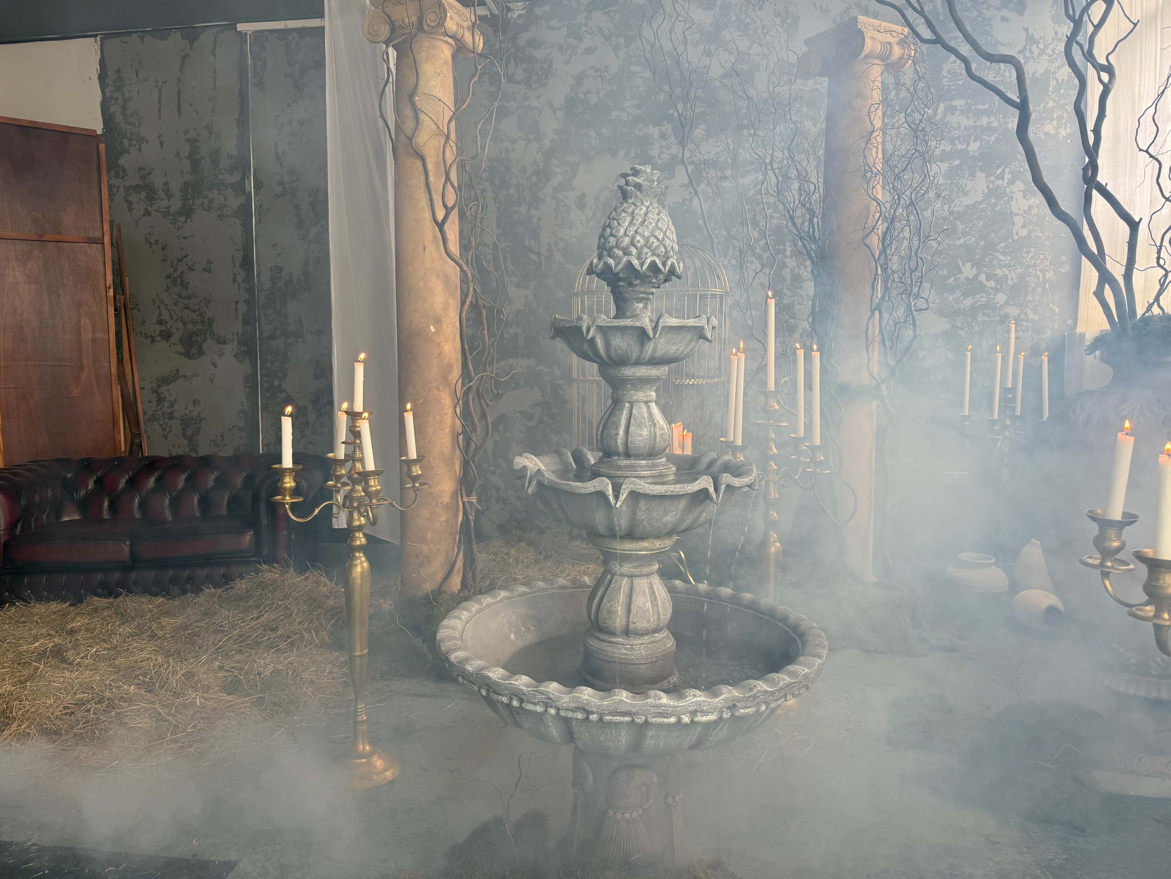 A decorative stone fountain with a pineapple sculpture sits in a foggy room surrounded by candelabras and overgrown greenery.